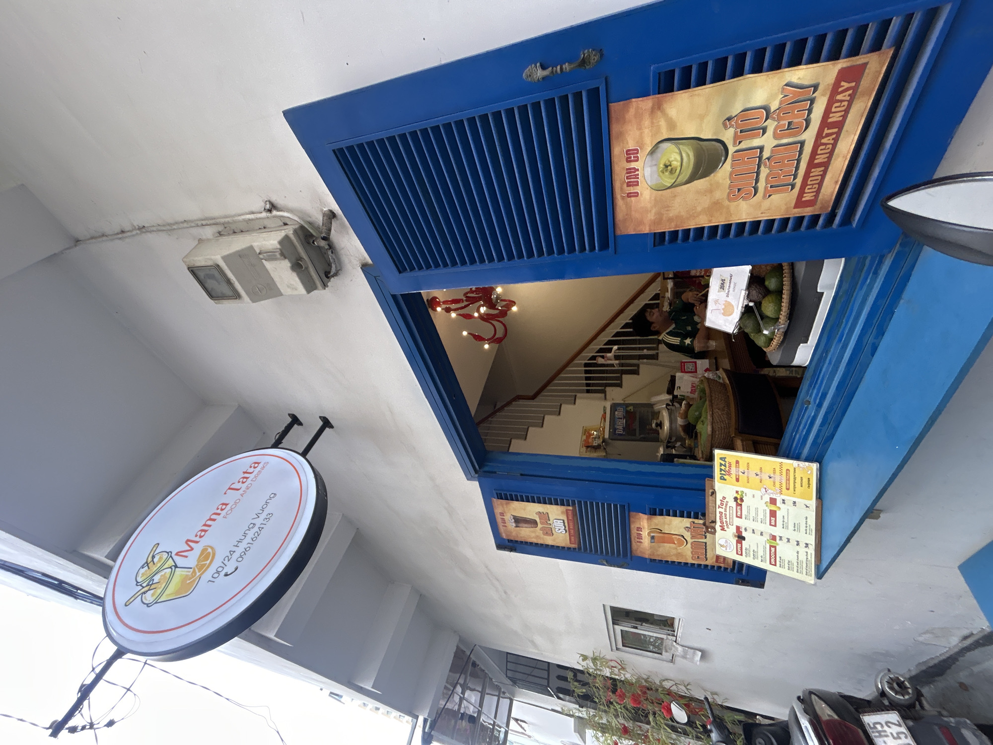 The image shows the exterior of "Mama Tata FOOD AND DRINKS" at 100/24 Hung Vuong in Nha Trang, Vietnam. An open, blue window or door serves as the service area. Posters and menu cards advertising drinks and food items such as "Sinh To Trai Cay" and pizza are attached to the blue frame. Inside the shop, a person stands near a counter with avocados, and there is a stairway leading upwards. A round sign displaying the shop's name and contact information hangs on the building's exterior. A motorbike is parked in front of the building.