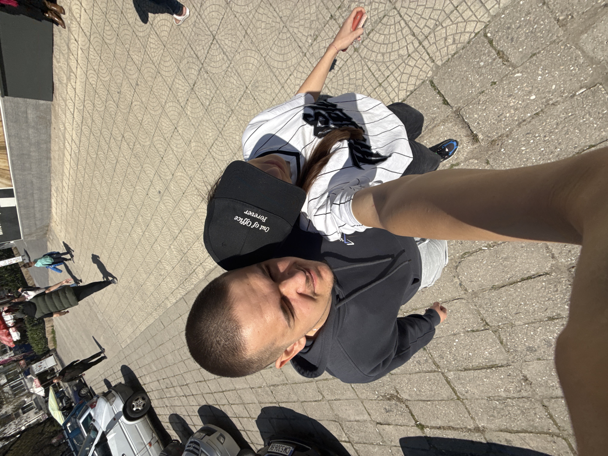 A selfie taken in Plovdiv, Bulgaria, shows a man in a dark hoodie standing next to a woman wearing a baseball cap and a white pinstripe shirt. The background includes a pedestrian area paved with geometric pattern stones, other pedestrians in the distance, and parked cars along a street.