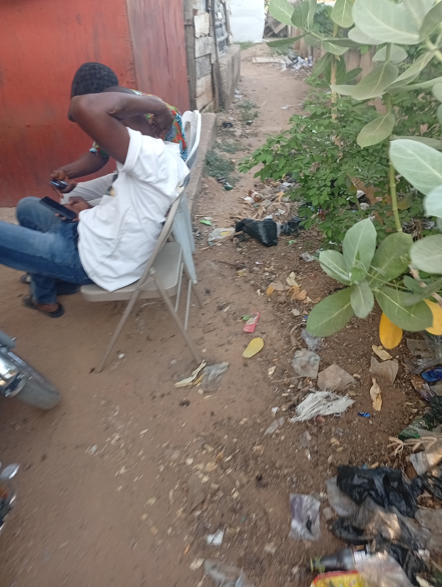 Two dark-skinned males are sitting on white plastic folding chairs outdoors. The foreground and midground are littered with trash. In the lower left corner, part of a motorcycle is visible. The location is in the city of Adankolo, Nigeria.