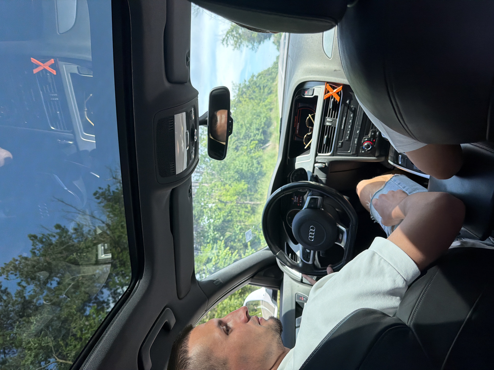 The image shows the interior of a gray Audi car, likely an A4 or A5 model, driving on a road near Obretenik, Bulgaria based on the exterior trees. The driver, a light-skinned man with short dark hair and a beard, is looking forward. The passenger's legs are visible. An orange "X" shaped object is on the dashboard, with sunglasses near it. The car has a sunroof. The rearview mirror is positioned in the center of the windshield. The steering wheel is an Audi brand with a central logo. The environment suggests a daytime setting with green foliage outside.