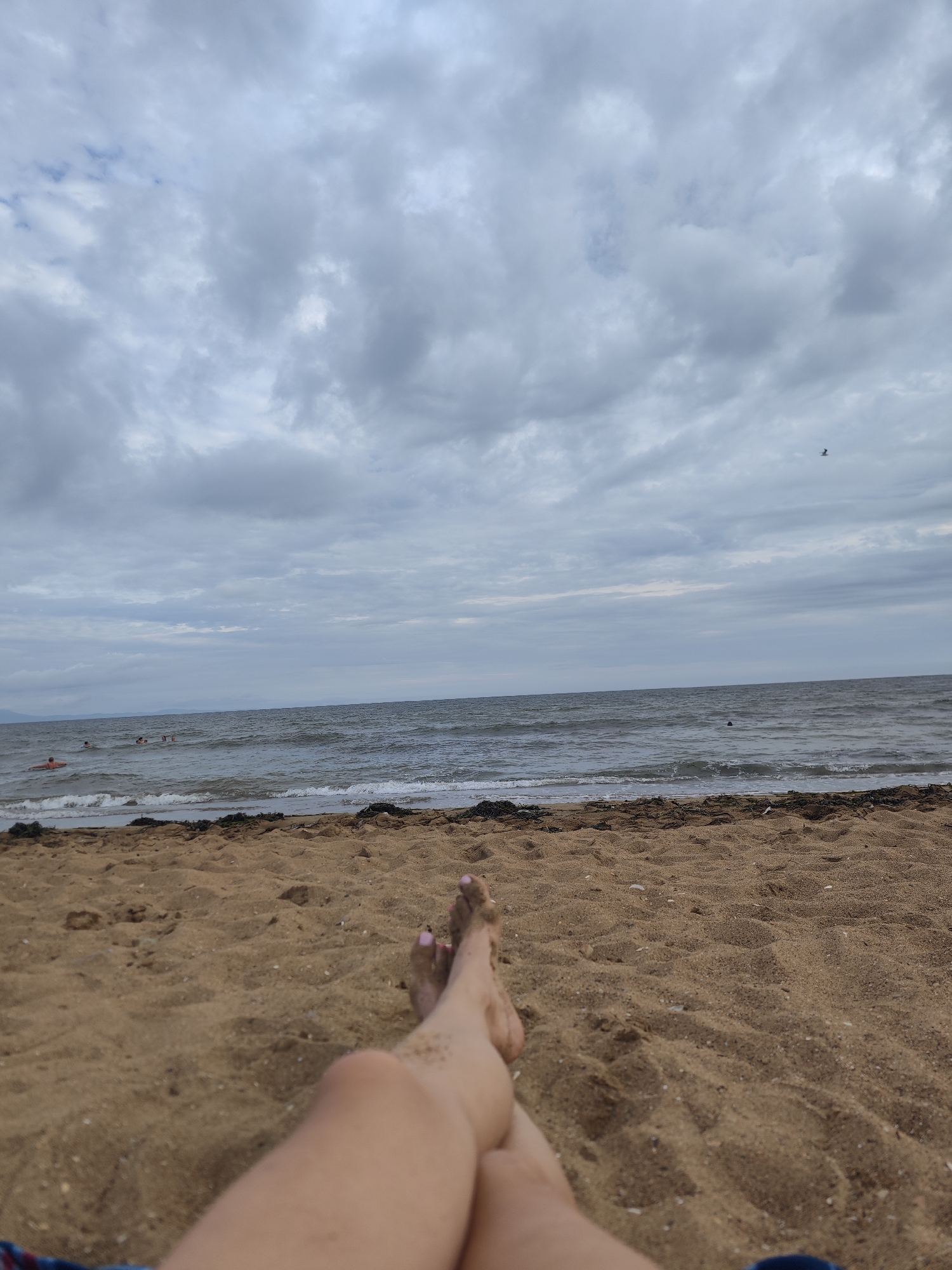 The image shows a first-person perspective of someone relaxing on a sandy beach. The foreground is dominated by the person's legs and feet, which are covered in sand. The feet have painted toenails, suggesting a leisurely activity. Beyond the sand, the sea is visible, with moderate waves and a few people swimming or wading in the water in the distance. The sky is overcast with a thick layer of clouds, giving the scene a subdued, perhaps cooler, atmosphere. A single bird is captured in flight against the cloudy sky. The overall impression is one of a quiet, possibly a bit gloomy, day at the beach. There is no information in the image to suggest the location is Vladivostok, Russia.
