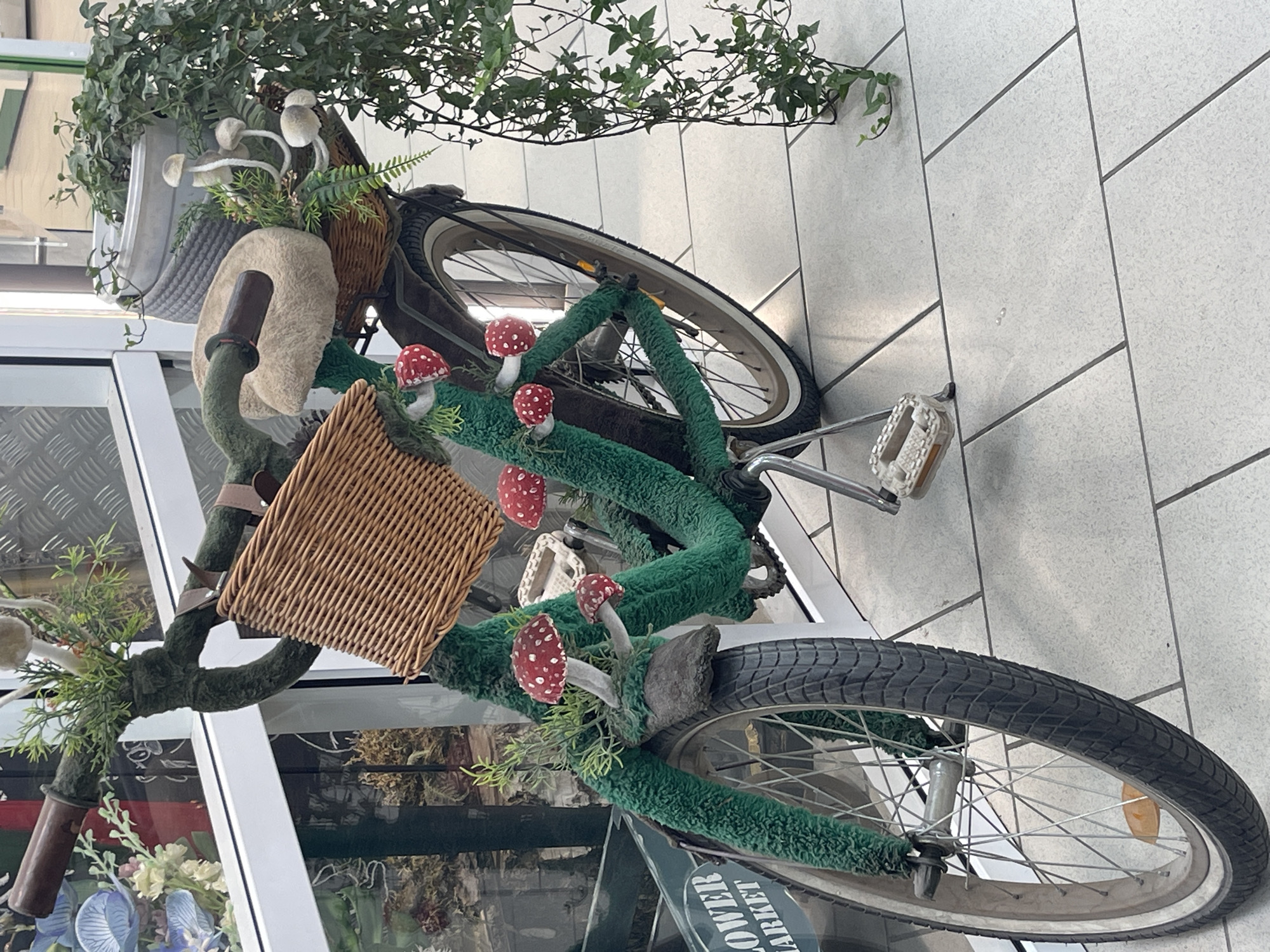 This is a photograph of a bicycle that has been decorated with artificial foliage and toadstool ornaments. The bicycle is leaning against a tiled wall, with one wheel resting on the ground. The front of the bicycle is adorned with green fuzzy material that resembles moss or grass, with several red and white toadstool decorations placed on it. A small wicker basket is attached to the handlebars, and a larger basket is visible behind the seat. A potted plant with green leaves is also placed on the front of the bicycle. The scene appears to be outdoors, possibly in a public space given the tiled flooring and the presence of what looks like shop windows in the background. The lighting suggests it is daytime. There is no visible text in the image. The overall impression is whimsical and decorative.