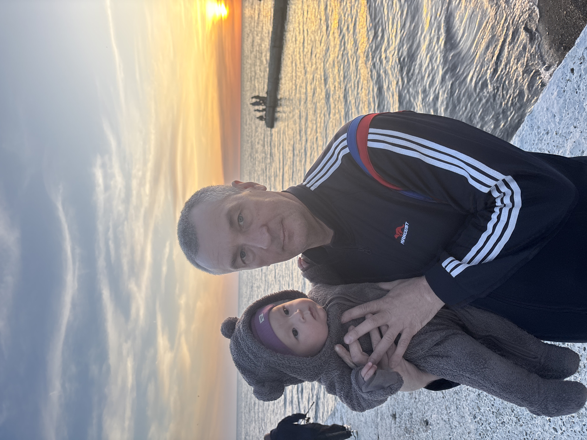 A man in a dark tracksuit is holding a baby in a hooded onesie, on a concrete surface next to the sea. The tracksuit has three white stripes on the sleeves. The baby is wearing a purple hat under the hood. The sea is to the right, with a breakwater structure visible in the distance. The sky is a gradient of blue and orange, suggesting either sunrise or sunset. The scene appears to be in Sochi, Russia due to the specific architectural details of the breakwater.