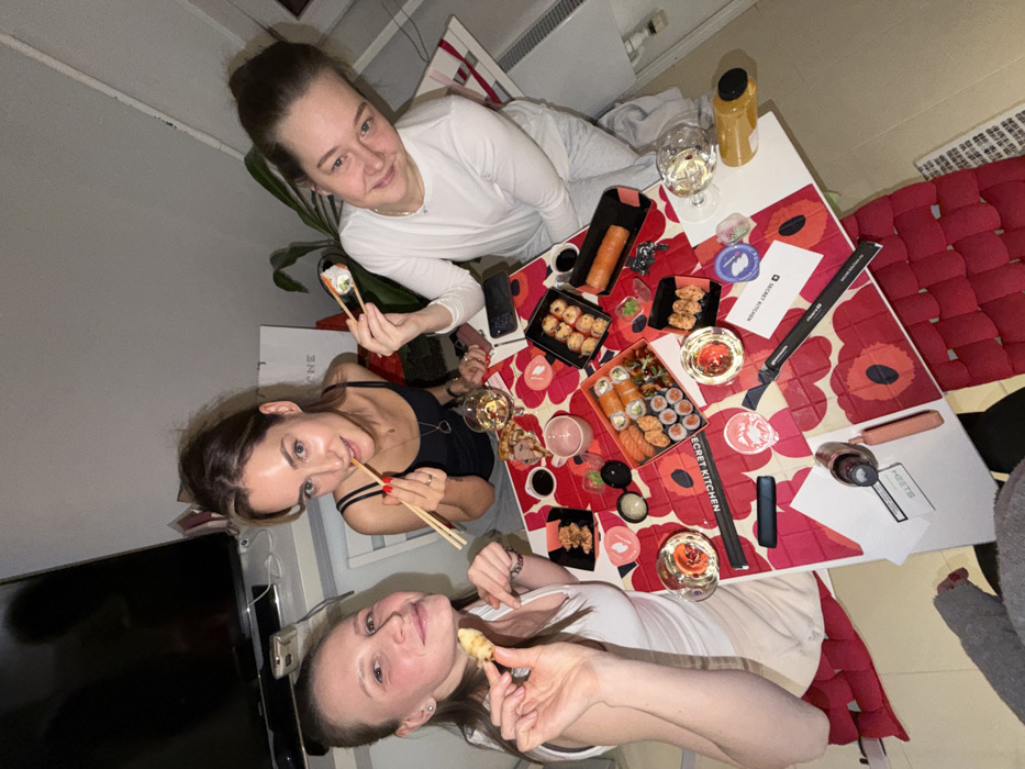 Three women are gathered around a table, enjoying a sushi meal. The table is covered with a floral tablecloth and various sushi platters, along with beverages like wine and juice. Chopsticks are in use, and the women appear to be socializing and smiling. A television and a red woven seat are visible in the background. The image is taken from a slightly elevated angle, giving an overhead view of the scene.