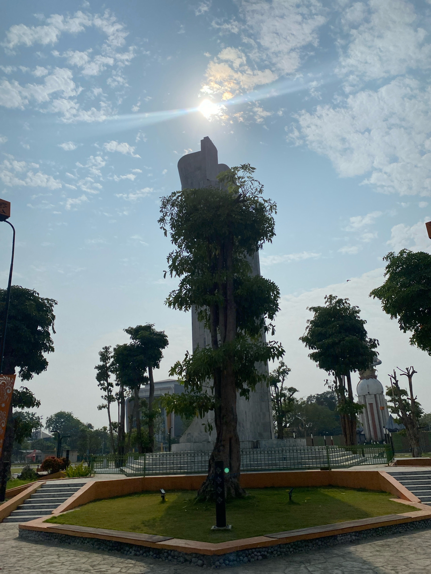The image depicts a view of the Monument Dwi Darmo in Sidoarjo, Indonesia, on a clear day with a bright sun and scattered clouds. A tall, imposing monument, possibly made of concrete or stone, dominates the center of the frame, partially obscured by the foliage of a large tree in front of it. The monument has a distinctive, abstract shape. Surrounding the monument are several other trees, some appearing taller and more slender than the central one, and some that have been pruned.
In the foreground, there is a paved area and a meticulously manicured lawn bordered by an orange-colored structure. Steps lead up to this lawn from the paved area. A wooden bench is visible on the left side of the lawn. Behind the central monument and trees, other structures can be seen, including a white, cylindrical tower-like building with a conical top, and further in the background, what appears to be a modern building with glass elements. A metal fence encloses a portion of the monument area. The overall scene suggests a public park or memorial site.