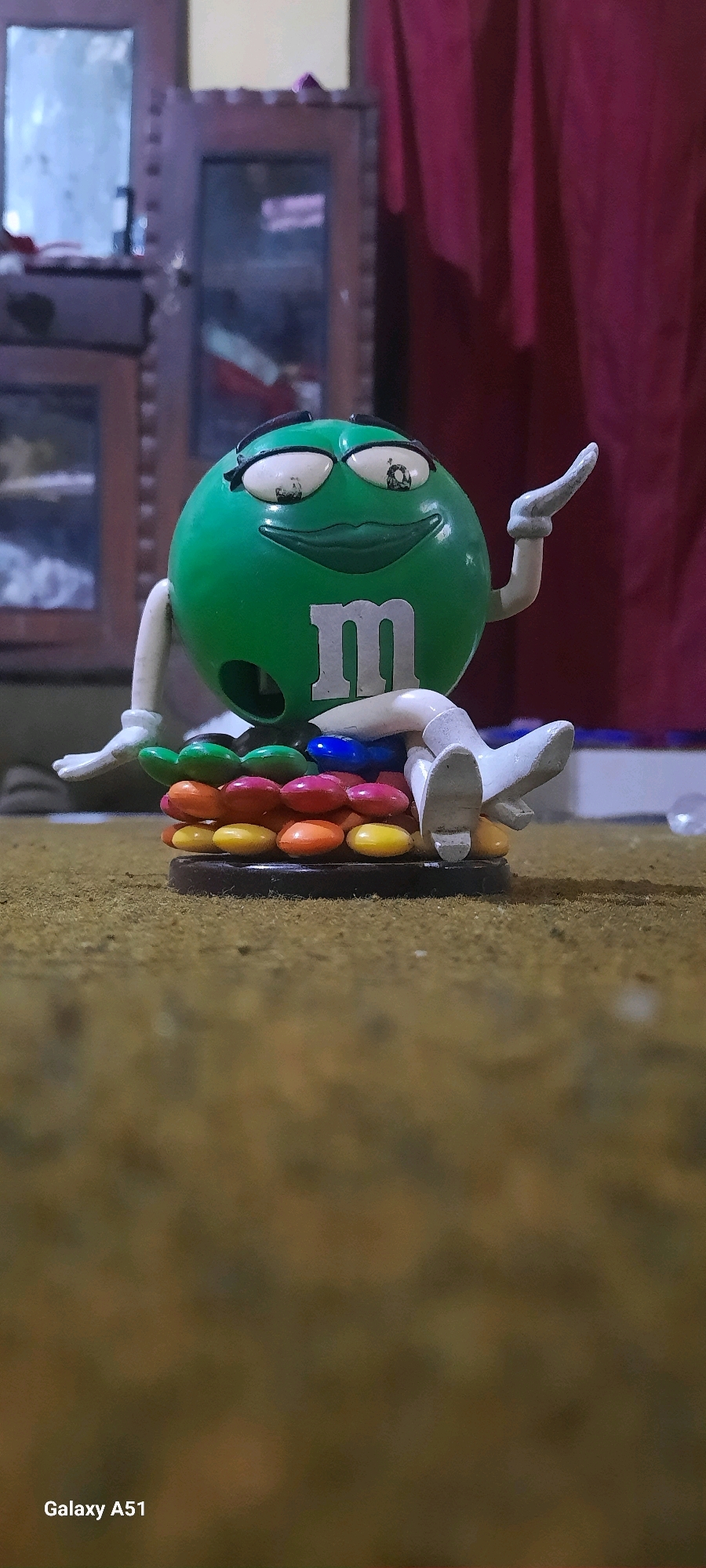 A plastic green M&M character figurine with white limbs and a white letter "m" on its chest is sitting on a layered base of colorful candies. The figurine has exaggerated facial features, including large white eyes with black pupils and eyelashes, and large red lips. Its arms are bent, with the left arm resting on its lap and the right arm raised with the hand open. Its legs are bent and crossed, with white boots. The candy base consists of multiple rows of round candies in shades of red, orange, yellow, blue, and green. The figurine and candy base are placed on a brown, textured surface that appears to be carpet or a similar material. In the background, a blurry wooden cabinet with glass doors is visible, and a dark red curtain hangs on the right side. A portion of a mirror reflecting light is visible on the cabinet. The overall lighting is dim. The bottom left corner of the image displays "Galaxy A51".