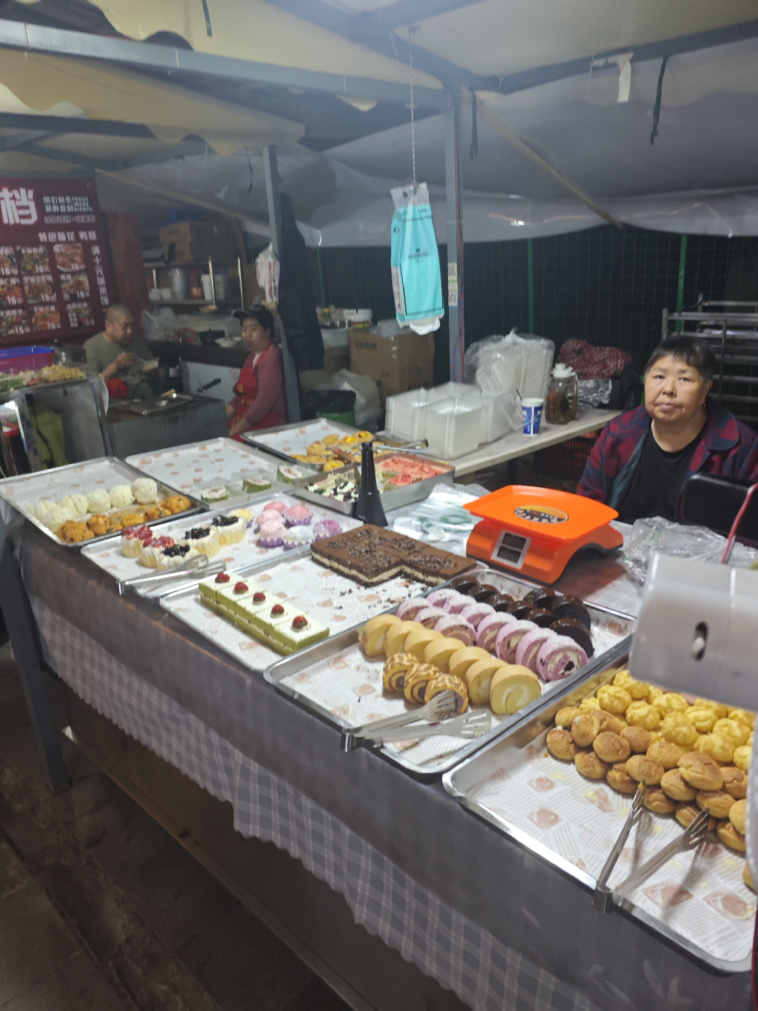 The image shows a street food stall in Shapingba District, China. An Asian woman stands behind a table covered with trays of various desserts, including cakes, pastries, and cream puffs. An orange scale sits on the table next to her. Further back, two other people are working at a cooking station. A menu board is visible in the background. The stall is under a makeshift tent.