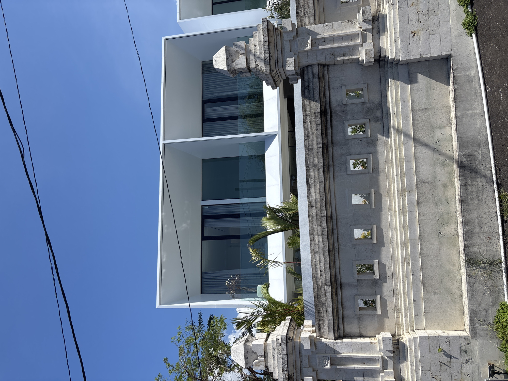 A low-angle shot shows a modern white building with large windows next to a traditional Balinese stone wall. The building has a boxy design, contrasting with the ornate carvings of the wall. Tropical plants are visible between the two structures. The sky is clear and blue. The location is Kuta Selatan, Indonesia. Power lines stretch across the upper left of the image. A sidewalk is visible on the right.