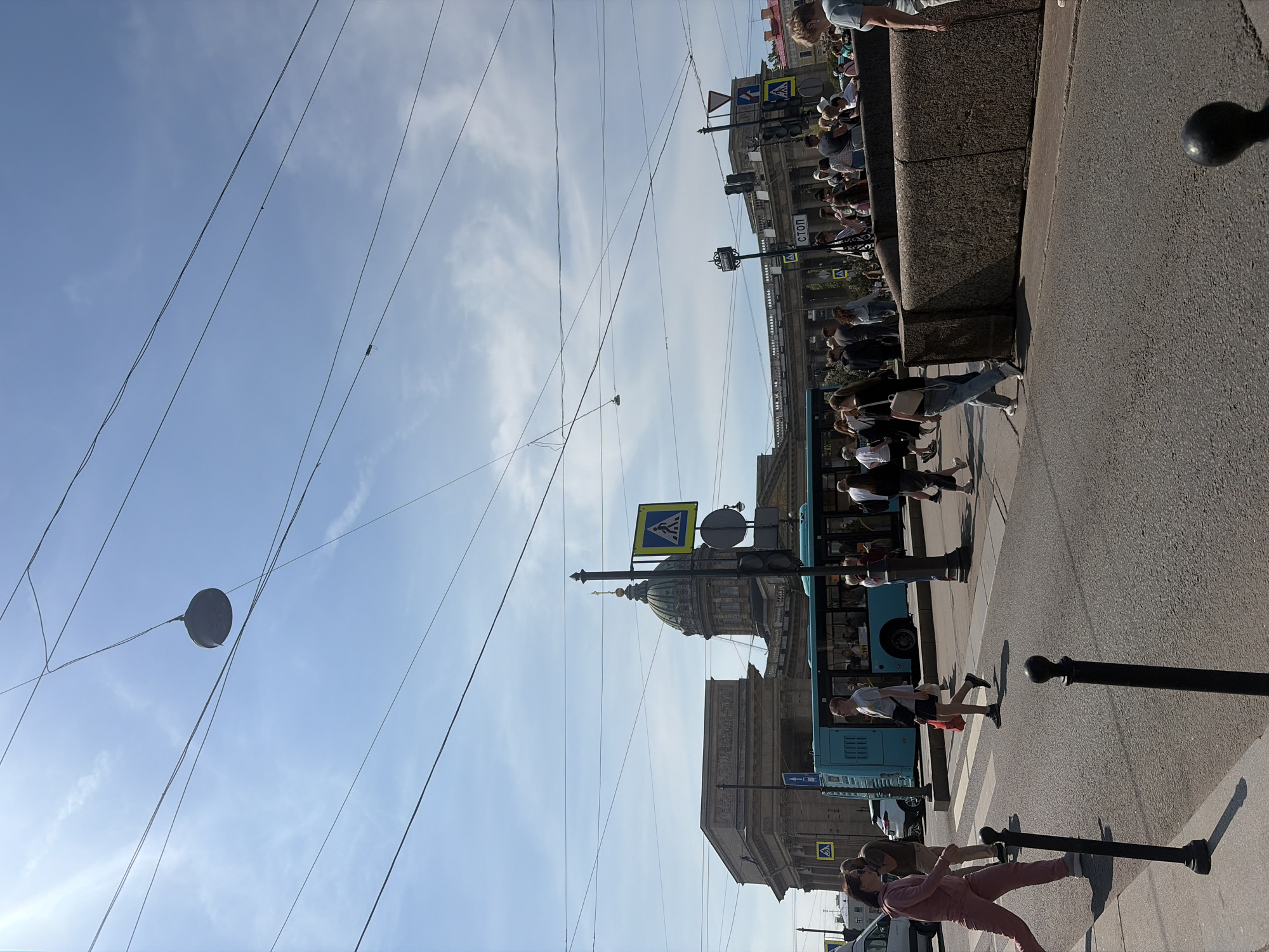 The image depicts a street scene in Saint Petersburg, Russia, likely in front of Kazan Cathedral, recognizable by its distinctive dome and colonnade. A bright blue bus is stopped at a crosswalk, with several pedestrians crossing the street. The sky is clear with a few wispy clouds, and overhead, numerous electrical wires crisscross the frame, a common feature in urban environments. On the left, a woman in a pink tracksuit is walking across the street, while a man in a white t-shirt and dark shorts walks in the same direction. Behind them, other pedestrians are visible, some standing near the bus and others walking. A white SUV is parked on the left side of the street. Traffic lights and pedestrian crossing signs are present. The buildings in the background showcase neoclassical architecture. The scene appears to be a busy urban setting on a sunny day.