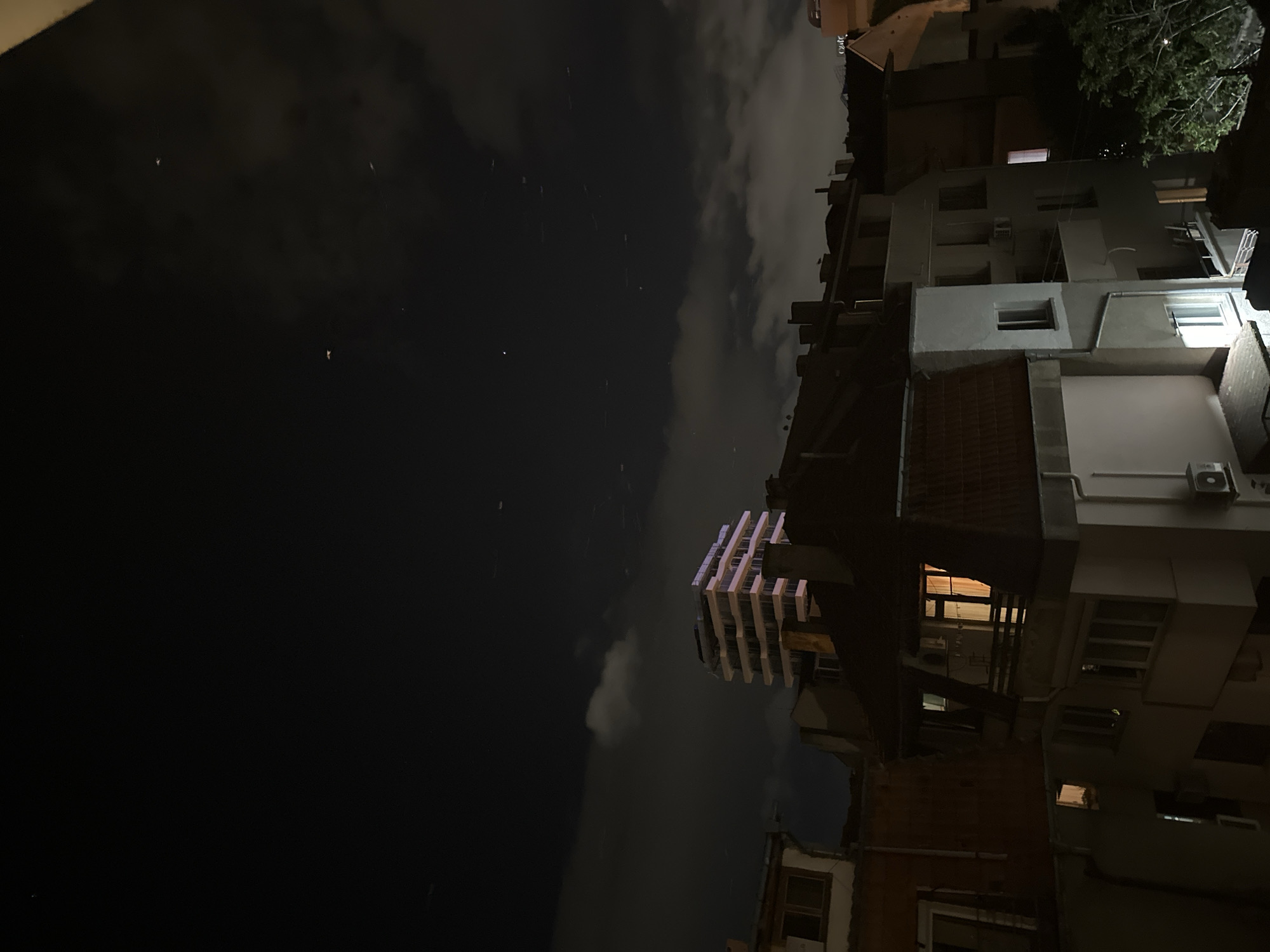 A nighttime view of buildings in Varna, Bulgaria. The sky is dark with clouds and a few visible stars. Buildings of varying heights and architectural styles are visible. One building has illuminated floors. Some buildings have balconies. One building is of brick construction. Air conditioning units are visible on some buildings. Trees are at the top right of the image.