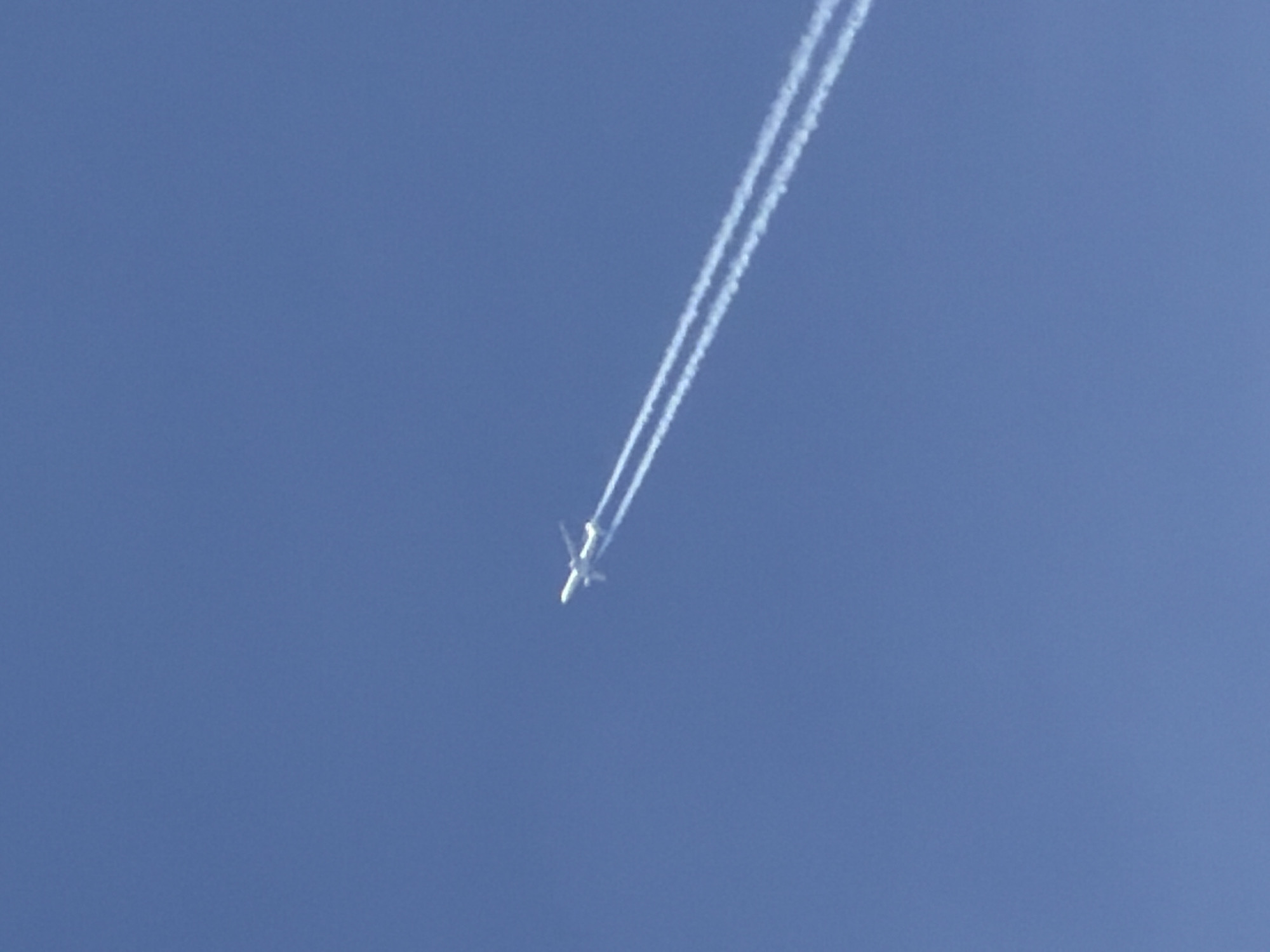 A daytime shot shows an airplane flying through a clear blue sky, leaving a long contrail behind. The airplane is small and white, and the contrail is thick and stretches from the top of the frame to just behind the plane. There are no visible buildings or other objects in the scene. The location cannot be determined from the image.