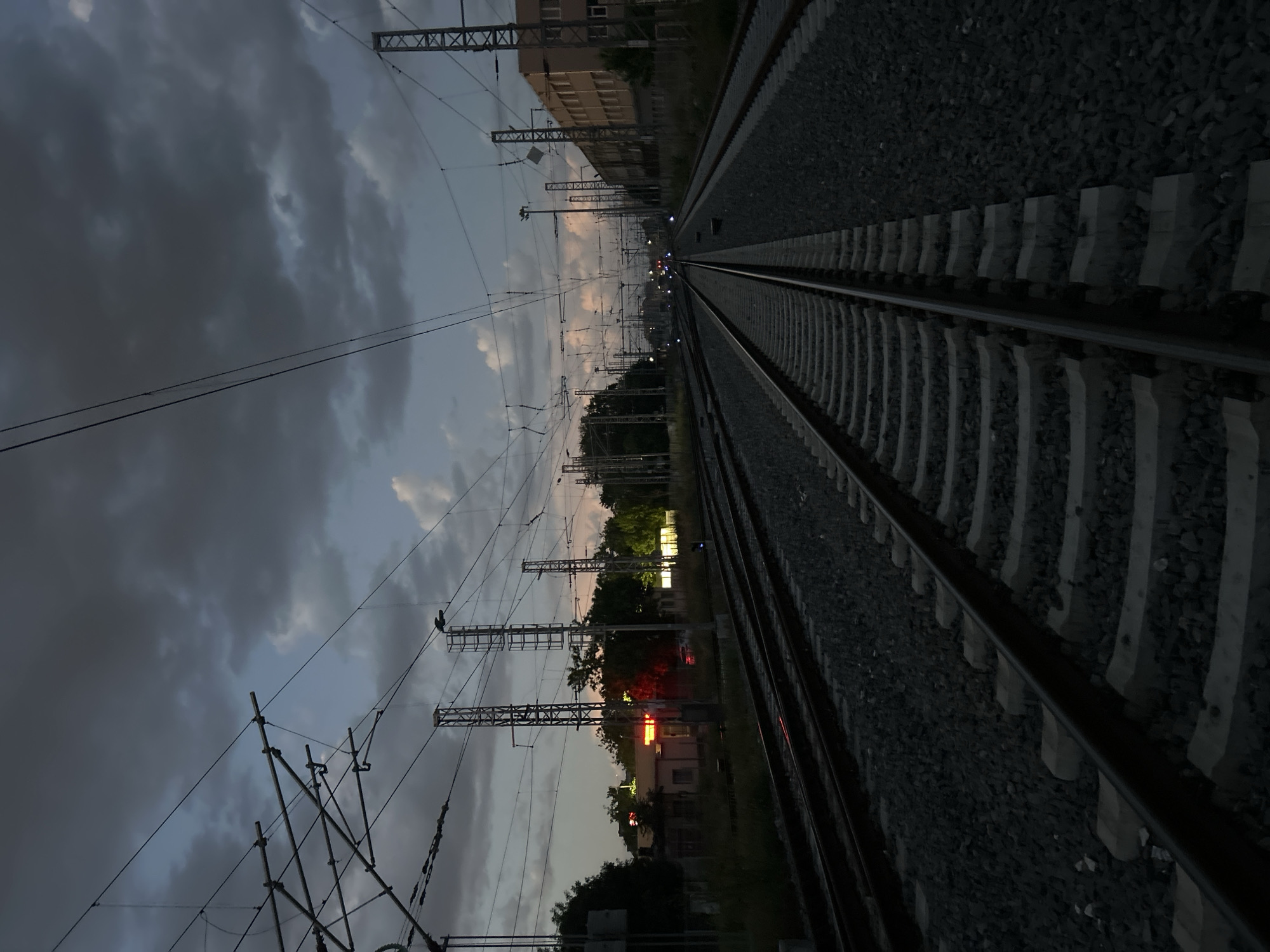 The image depicts a train track receding into the distance under a cloudy, twilight sky. The track is bordered by gravel and concrete sleepers. Overhead power lines and support structures stretch along the length of the tracks. In the middle ground, there are buildings and trees, with some lights visible. The scene is likely near the railway station in Plovdiv, Bulgaria, given the presence of train tracks and associated infrastructure. There are no people visible in the image. The overall impression is of a quiet, industrial landscape at dusk.