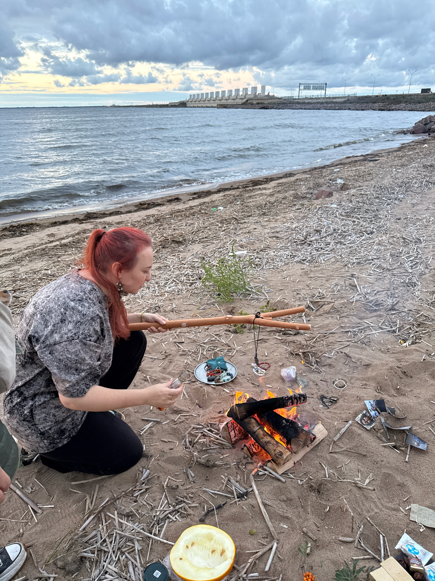 A woman with red hair pulled into a ponytail kneels on a sandy beach by a small bonfire. She wears a grey and black speckled shirt and black pants. In her right hand, she holds a small glass of amber liquid. In front of her, a fire burns with logs stacked in a metal grate. Scattered around the fire are various objects, including a plate with jewelry and crystals, a half-cut melon, and a few packets of candy. In the background, a body of water stretches to the horizon, with a large concrete structure, possibly a dam or industrial facility, visible in the distance. The sky is overcast with dramatic clouds. The overall setting appears to be a recreational area along the coast of Lomonosov, Russia.