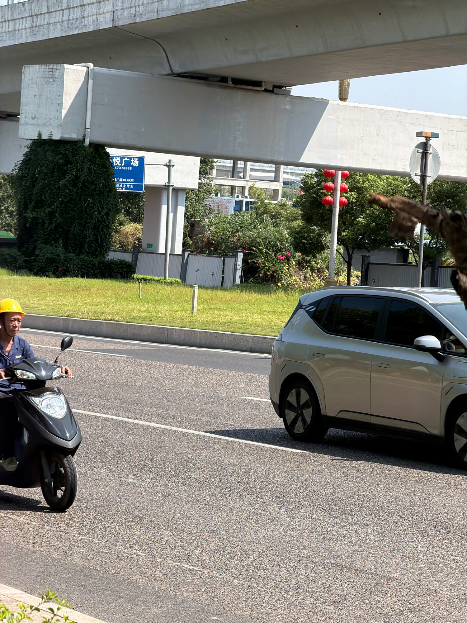 The image depicts a street scene in Jiangjin, China. In the foreground, a person wearing a yellow hard hat and a blue uniform rides a black scooter. To the right, a silver electric car is visible on the road. Behind them, a grassy embankment lines the street, leading up to an elevated highway overpass. A blue sign with Chinese characters and numbers is mounted on a support column under the overpass. Red lanterns hang from a pole in the distance, and trees and buildings are visible in the background. The sky is clear and blue.