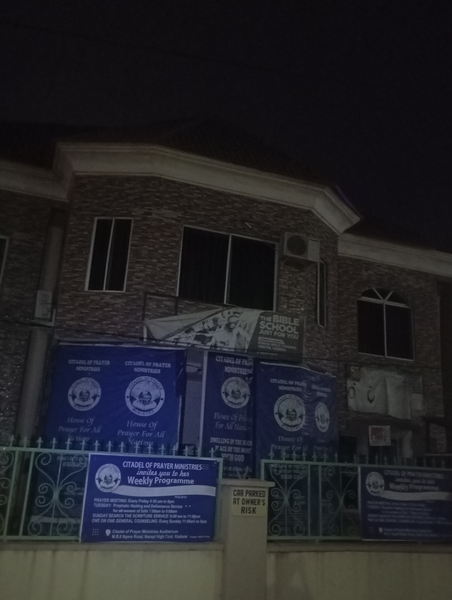 The image depicts the exterior of a building at night, likely a church or ministry called "Citadel of Prayer Ministries" located in Narayi, Nigeria. The building has a brick facade with multiple windows, some of which are dark, indicating they are not illuminated from within. An air conditioning unit is visible on the upper part of the building.
In front of the building, there are several large blue banners with white lettering. These banners appear to announce the ministry's name and some of its services, such as "House Of Prayer For All Nations". One banner specifically advertises "THE BIBLE SCHOOL JUST FOR YOU." Another banner, in blue and white, lists the "Weekly Programme" for Citadel of Prayer Ministries, detailing prayer meetings, prophetic healing, deliverance services, and scripture searches, with specific timings and days. The address is listed as M.B.B.S Nyeré Road, Narayi High Cost, Kaduna.
A metal fence with decorative scrollwork separates the building from the foreground. A sign on a white pillar to the left of the fence reads "CAR PARKED AT OWNER'S RISK". The lighting in the image is dim, suggesting it was taken at night, with some artificial light illuminating the banners and building facade. The overall scene conveys a sense of a religious institution operating in a community setting.