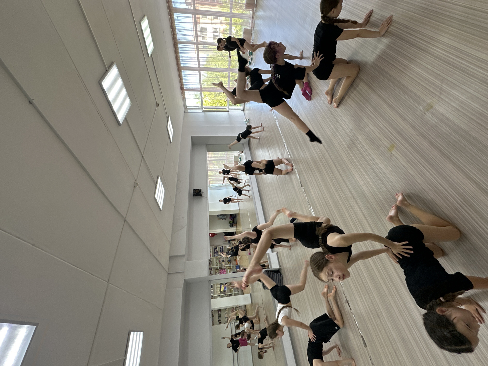 The image depicts an indoor scene of a dance or gymnastics class primarily involving young girls and teenagers in Votkinsk, Russia. They are dressed in black athletic wear, including shorts and t-shirts or leotards, with most being barefoot or wearing black socks.
The setting is a large, well-lit studio featuring a light-colored wooden floor and white walls. One wall is entirely mirrored, reflecting the participants and creating a sense of depth and a larger group. Large windows along one side of the studio allow abundant natural light to flood the space, suggesting it is daytime, likely sunny. The ceiling is equipped with multiple rectangular fluorescent light panels, adding to the bright illumination.
The participants are actively engaged in various stretching, posing, and balancing exercises. Several are on the floor, holding different dance or gymnastic positions, such as leg extensions, back arches, and kneeling stretches. In the foreground, one girl holds her leg high in an extended pose, supported by another. Their facial expressions convey concentration and effort as they execute the movements. The overall atmosphere suggests a focused training session.
A notable detail is a partially visible logo or text, possibly "КИ" or "ХИ" in Cyrillic, on the black t-shirt of one of the girls in the mid-ground. The photograph is taken from an elevated and tilted perspective, adding a dynamic feel to the active scene.