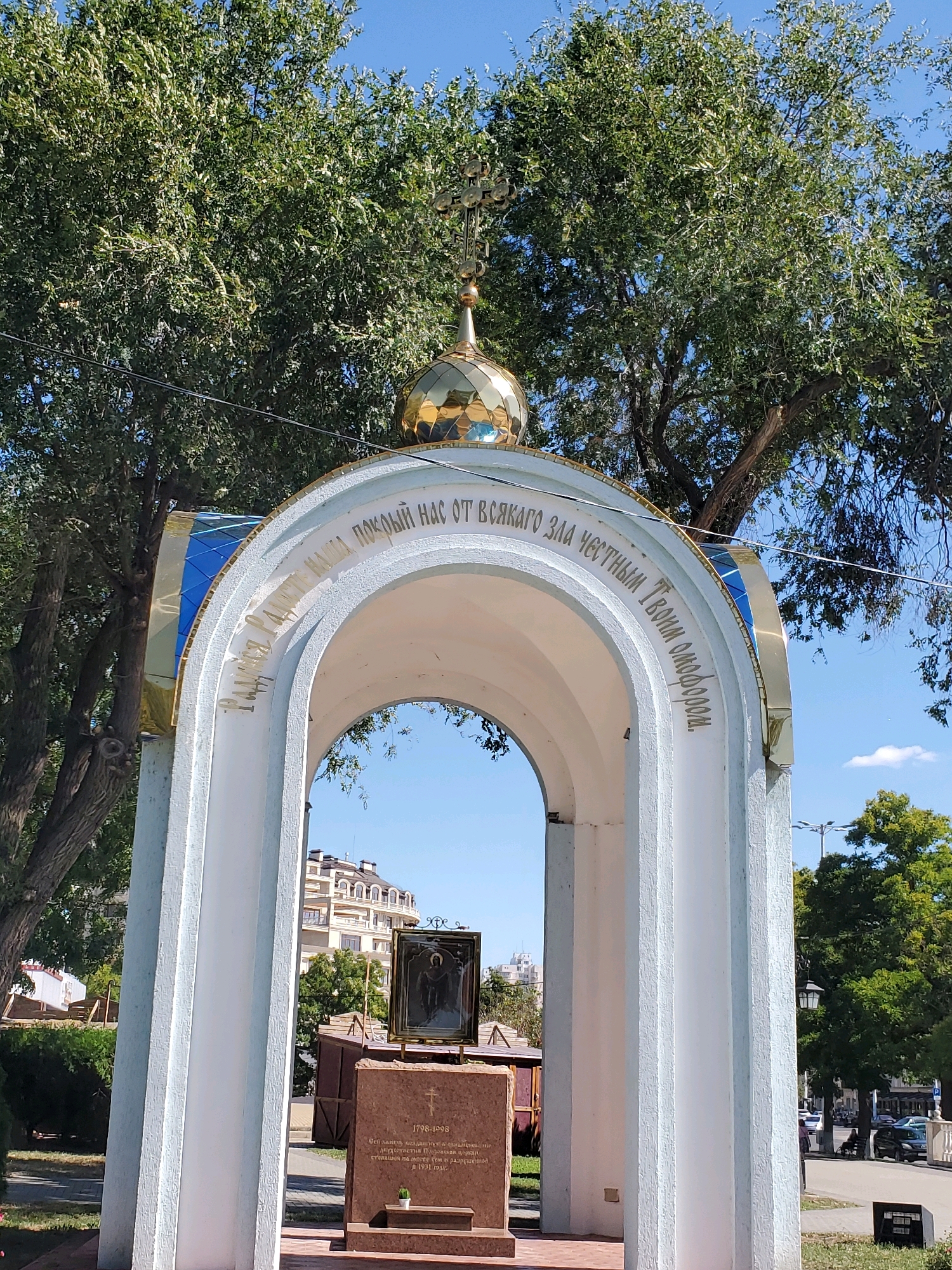 The image shows a white arched structure topped with a golden dome and a cross. Golden lettering is inscribed on the arch, which reads "Радуйся, Радосте наша, Покой наш от всякаго зла честным Твоим Омофором." This translates to "Rejoice, our Joy, our Peace from every evil with Your honorable Omophorion."
Beneath the arch, a memorial plaque is visible, featuring an icon of a saint in a golden frame. The plaque itself is made of reddish-brown stone with an inscription below the icon. The inscription appears to be in Russian and includes dates "1798-1998". A small potted plant sits on a ledge below the inscription.
The memorial is located in a park-like setting with mature trees providing shade. In the background, there are buildings, streetlights, and a street with cars and a few people. The sky is clear and blue. The memorial is likely part of a religious site or a commemorative monument. The context provided states the location is Tiraspol, Moldova.