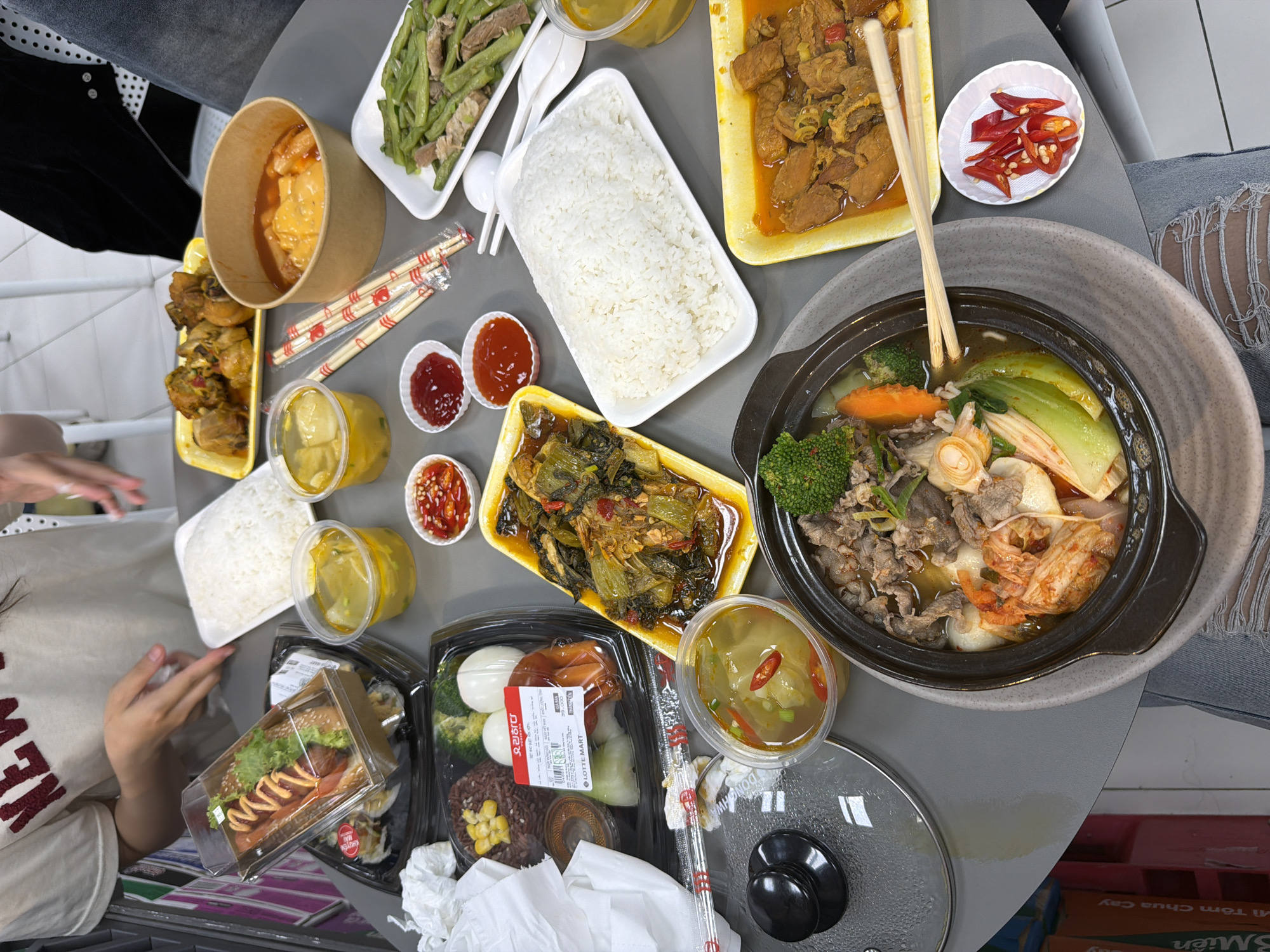 This is an overhead shot of a table laden with various takeout dishes, likely taken indoors. The arrangement suggests a shared meal. The presence of "LOTTE MART" labeling on a package suggests the scene may be in Đà Nẵng, Vietnam, where Lotte Mart has locations.
Visible on the table are multiple containers holding different foods. A dark clay pot-style bowl contains what appears to be a stew or soup with visible vegetables, meat, and noodles, with chopsticks resting on top. Several white rectangular containers hold what looks like plain white rice. Other containers hold an assortment of dishes, including what might be marinated meats, braised greens, and a sandwich. There are also small containers of sauces and sliced chilies. Some of the containers are clear plastic, allowing the food inside to be easily seen, while others are paper.
Two people are partially visible in the frame, with parts of their clothing and hands showing. One person wears a beige shirt with the word "NEW" printed on it, and another is in a black jacket. The presence of food delivery-style containers and takeout bags implies the meal was sourced from various establishments and assembled at this location. A lid from one of the containers also rests on the table. Overall, the scene depicts a casual dining setup with a variety of different cuisines.