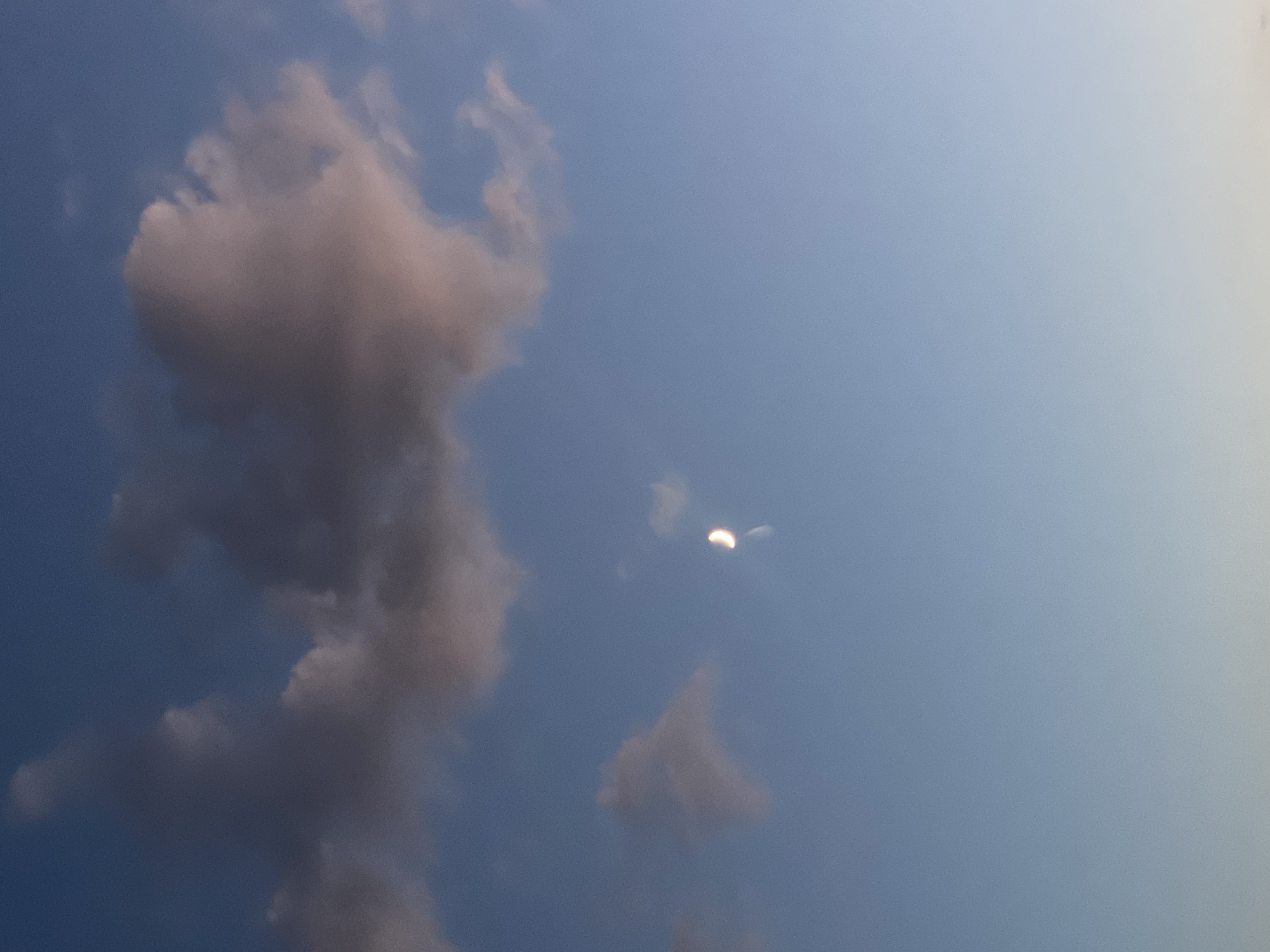 The image captures a serene sky scene above Novi Petrivtsi, Ukraine. A crescent moon is visible in the center-right of the frame, appearing as a thin, luminous sliver against the soft blue expanse. Several clouds are scattered across the sky; a large, dark cloud formation dominates the upper left, with lighter, pinkish hues suggesting it might be illuminated by the setting or rising sun. Another smaller cloud is positioned to the left of the moon, appearing translucent. The overall atmosphere is tranquil, indicative of either dawn or dusk. The environment is solely the sky, with no ground elements or human presence discernible in the frame.