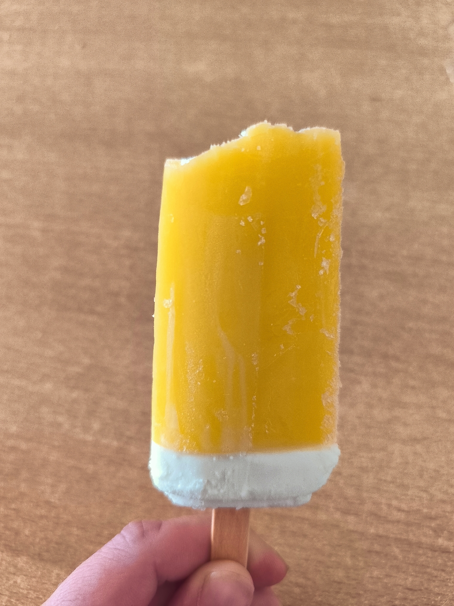 A close-up, eye-level shot captures a person's hand holding a half-eaten, yellow popsicle with a white bottom. The popsicle has a bite taken out of its top, revealing a slightly rough texture. The background is a textured, light brown surface, resembling wood. The scene is likely indoors, given the lighting. The exact location in Wervershoof, Netherlands cannot be determined from the visual information provided.