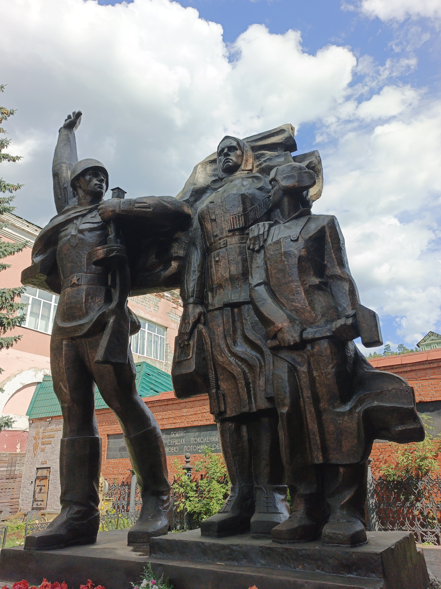 A bronze sculpture of three figures stands outdoors under a cloudy blue sky. The sculpture is located in Zlatoust, Russia. In the foreground, a statue of a male soldier in military uniform, wearing a helmet and boots, raises his left arm. He carries a rifle over his shoulder with his right arm. To his right, a male figure, also in military attire, stands with his hands clasped in front of him. A third figure, appearing to be female and dressed in civilian clothing, stands to the right of the male figures, with her hands clasped in front of her. Behind the figures, a two-story building with a pink facade and white-trimmed windows is visible, along with a green-tiled roof. Further back, a red brick building with a metal fence and green foliage is present. The base of the sculpture appears to be made of stone. Red flowers are visible in the very bottom of the image.