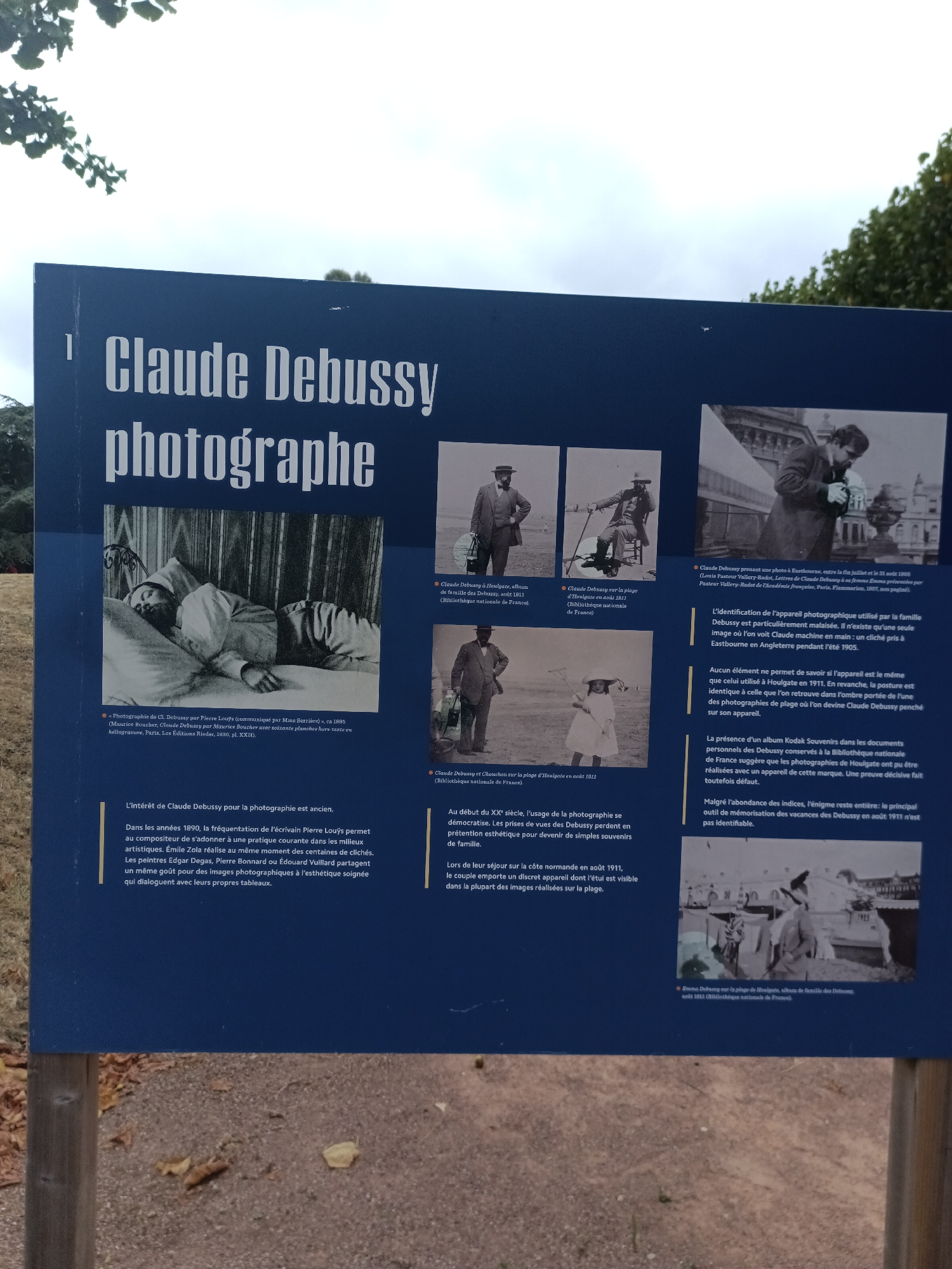 This image is a photograph of a historical marker or informational display in Houlgate, France. The display has a dark blue background and features text and several black and white photographs.
The main text at the top reads "Claude Debussy photographe." Below this are several captioned photographs and descriptive text.
One photograph shows Claude Debussy lying in bed, seemingly asleep, with a cap on his head. Another photograph shows a man identified as Claude Debussy on a beach with a cane, seated on a chair. A third photograph shows a child, possibly wearing a dress and hat, on a beach. A fourth photograph shows several people on a beach, one of whom is identified as Claude Debussy.
The text discusses Claude Debussy's interest in photography, mentioning his acquaintance with writer Pierre Louÿs and other artists who engaged in photography. It also describes the equipment used and some specific photographs taken by Debussy and his family. One caption mentions "Claude Debussy sur la plage d'Houlgate en août 1911," indicating a specific location and date within Houlgate, France. Another caption refers to "Claude Debussy et Chouchou sur la plage d'Houlgate en août 1911." The overall context is the amateur photography of Claude Debussy, with a focus on his visits to Houlgate.
The surrounding environment visible in the image is out of focus, but appears to be outdoors with trees and foliage, suggesting the marker is placed in a park or public area.