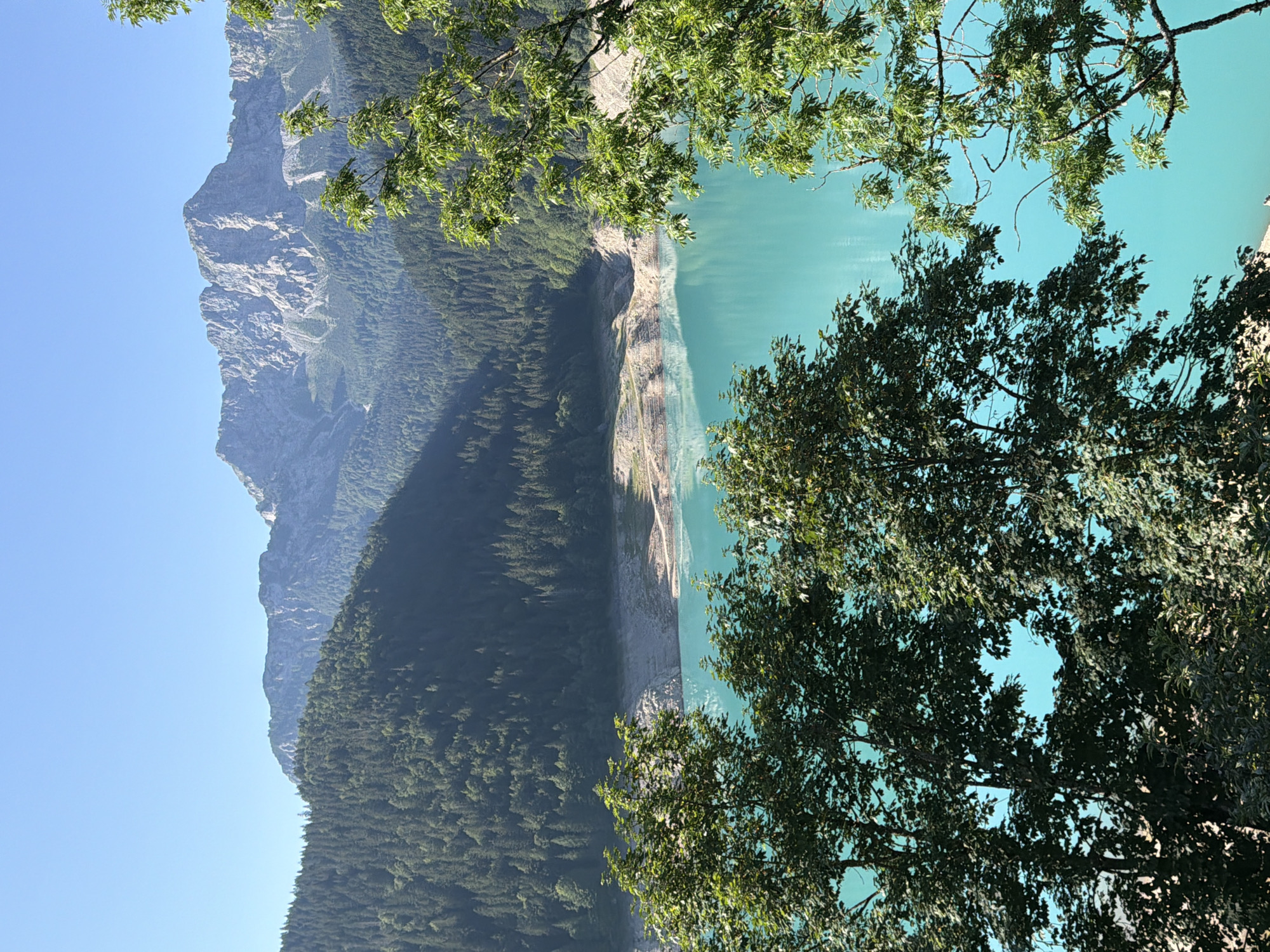An ultra-wide shot captures a serene mountain lake with turquoise waters, framed by lush green trees in the foreground. Majestic, rocky mountains rise in the background under a clear blue sky. The image, taken from an elevated viewpoint, shows the lake's shoreline with visible terraces and reflections on its still surface. The setting evokes a sense of natural beauty and tranquility.