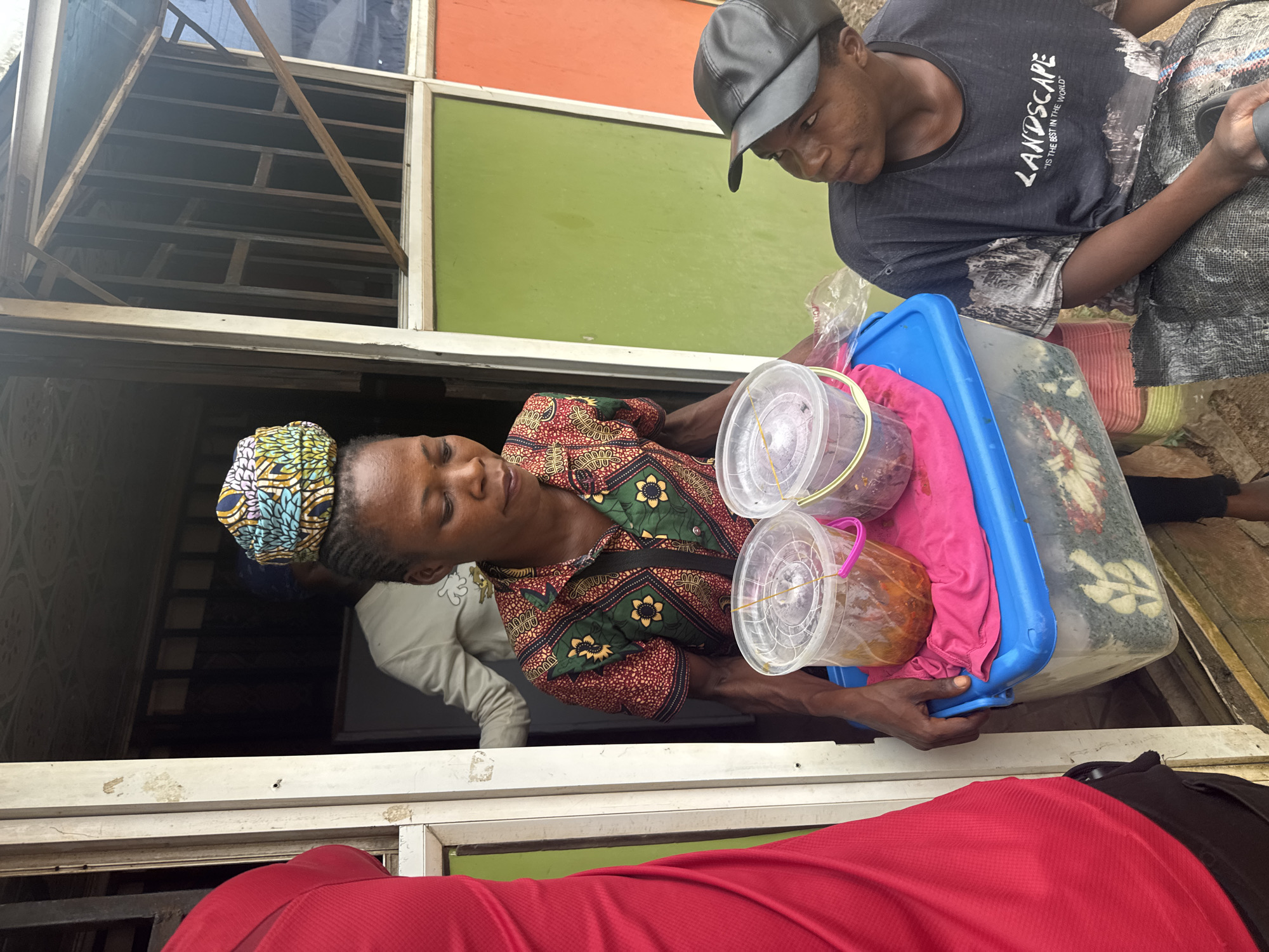 In Ihiagwa, Nigeria, an individual is carrying a clear plastic container filled with food items, possibly snacks or prepared meals, which is placed on top of a larger blue and clear plastic container. The larger container also holds food items. Both containers are topped with pink cloth. The individual is wearing a colorful, patterned shirt and a headwrap. To the right, another individual is seated, wearing a black t-shirt with "LANDSCAPE" printed on it and a black baseball cap. A third person, partially visible on the left, is wearing a bright red shirt. The scene appears to be outdoors, in front of a building with green and orange panels.