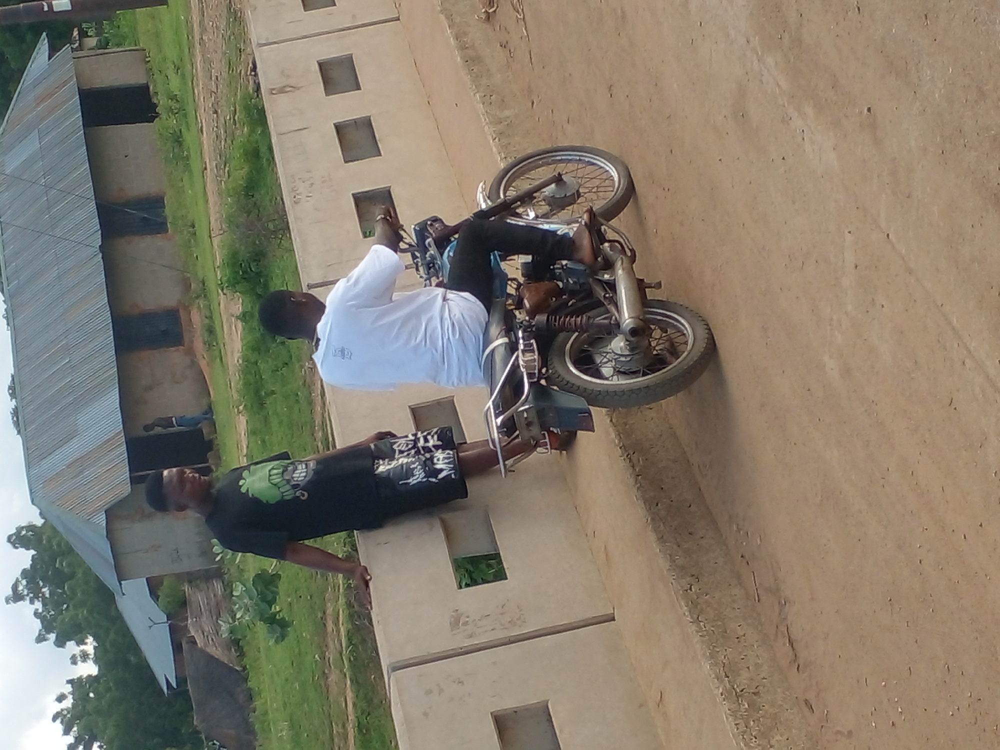 In the city of Unknown, Unknown, two young men are observed on a dirt road. One man, wearing a black t-shirt with a green graphic and black shorts with white lettering, leans against a concrete barrier. The other man, dressed in a white t-shirt and black pants, sits on a motorcycle. The motorcycle appears to be a standard model with a visible exhaust pipe and rear suspension. Behind them, several buildings with corrugated metal roofs and concrete walls are visible, nestled amongst lush green vegetation and trees. The overall setting suggests a rural or semi-urban environment. The lighting indicates it is daytime.