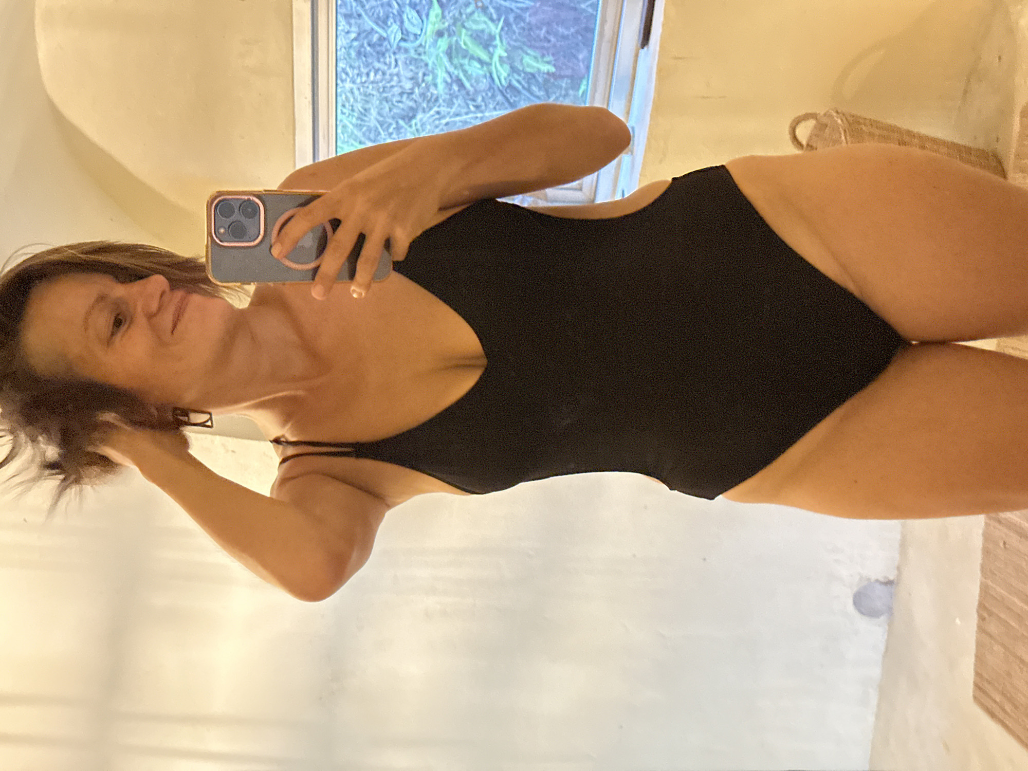 In a brightly lit bathroom with textured white walls and a window overlooking greenery, a woman with short brown hair and a playful expression is taking a selfie. She is wearing a black one-piece swimsuit with cutouts on the sides. Her left arm is raised, touching her hair, and she is holding a smartphone in her right hand, angled to capture her reflection in a mirror. The bathroom appears to be located in a tropical setting, possibly a resort or private residence, in the vicinity of Koh Phangan, Thailand, suggested by the window view and the presence of a woven basket in the background.