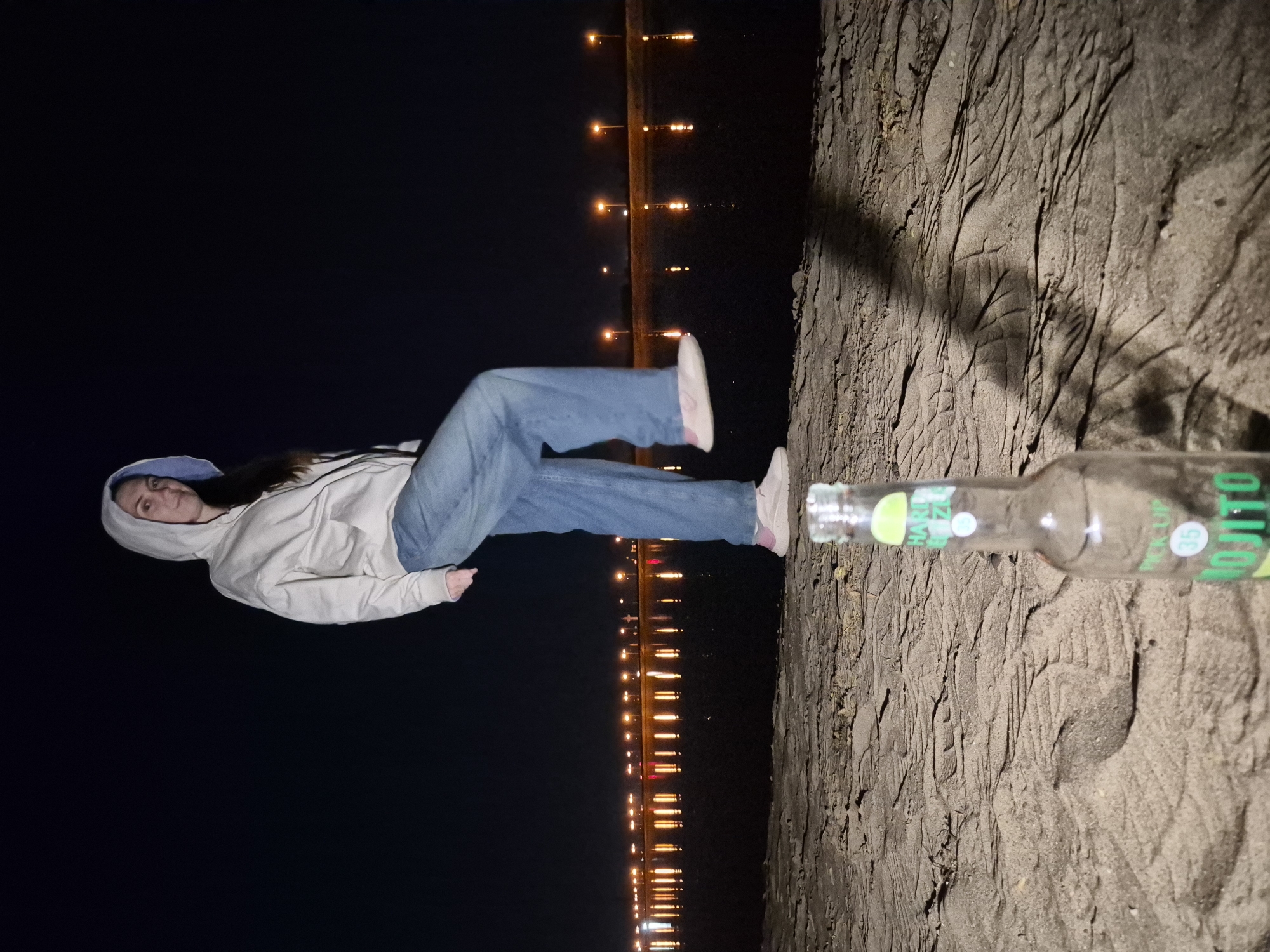 The image shows a woman in a light-colored hoodie and blue jeans, seemingly lying on a sandy surface. A partially visible glass bottle lies on the sand near her. In the background, a line of lights stretches along the horizon, suggesting a cityscape or shoreline at night. The location appears to be a beach or sandy area in the vicinity of a city, although the specific city is not identifiable from the image.