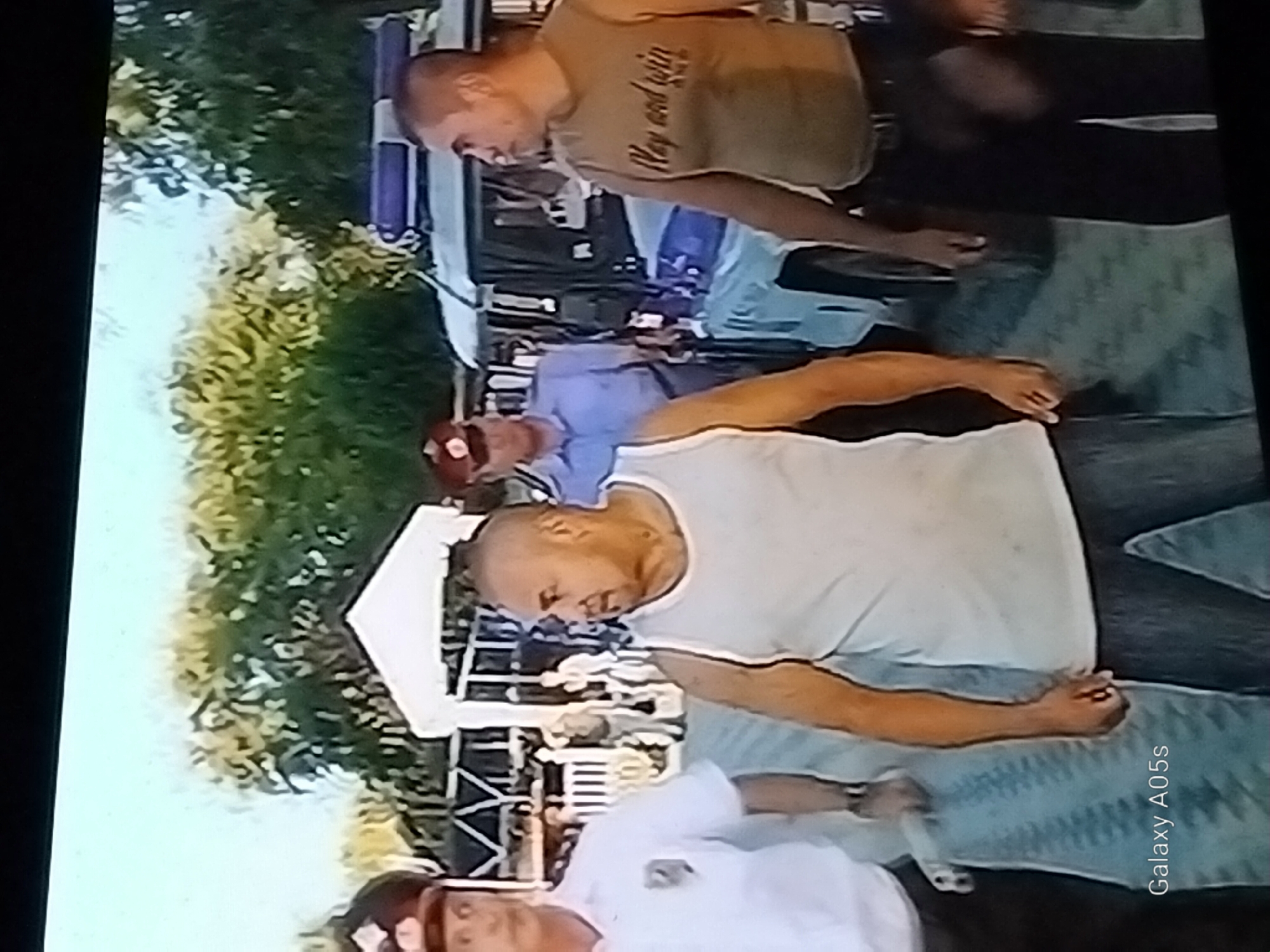The image shows a group of people outdoors, with a police vehicle visible in the background. Three men are in the foreground. The man on the left is wearing a white uniform with a badge, a red cap, and appears to be a police officer. He is looking forward. Next to him, a bald man in a white tank top and blue jeans is looking to his right. To the right of the bald man, another man in a sleeveless tan shirt with text on it, dark pants, and a belt is looking to his left. The text on his shirt appears to read "Play and win." Behind these men, another individual in a blue uniform and a red cap is standing near the police vehicle, which has a blue and white light bar on its roof. The environment appears to be a paved area with trees and a white tent-like structure in the background. The image is blurry and pixelated, suggesting it was taken from a video recording or a low-resolution photograph. The timestamp "Galaxy A05s" is visible in the bottom left corner. The location is identified as Druzhba, Russia.