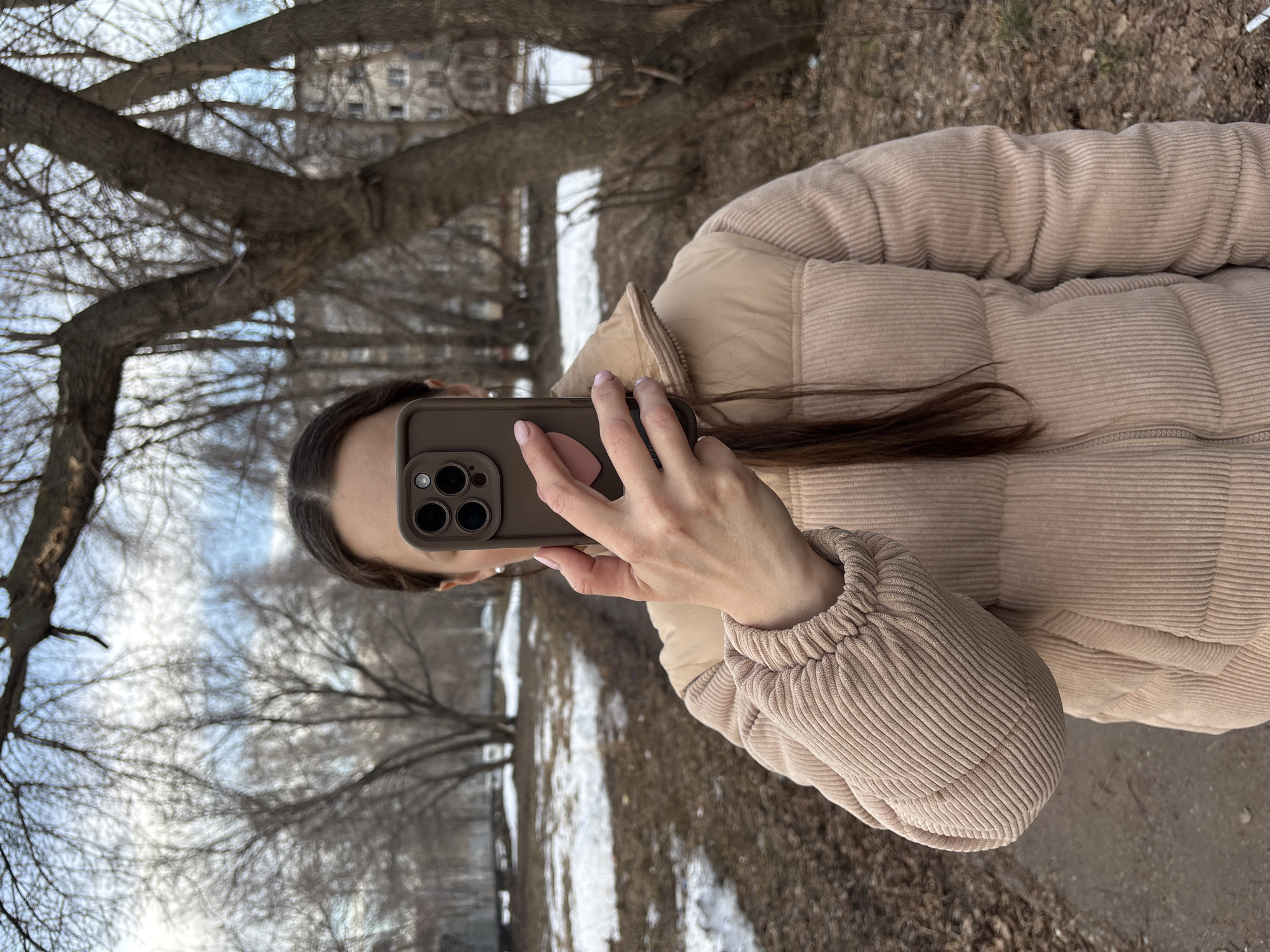 A woman is holding a smartphone in a brown case, presumably taking a selfie or a photograph. She is wearing a beige corduroy puffer jacket. The background shows a leafless urban park or wooded area with patches of snow on the ground. A building is faintly visible in the distance. The location is likely Ulyanovsk, Russia, based on the user's prompt.