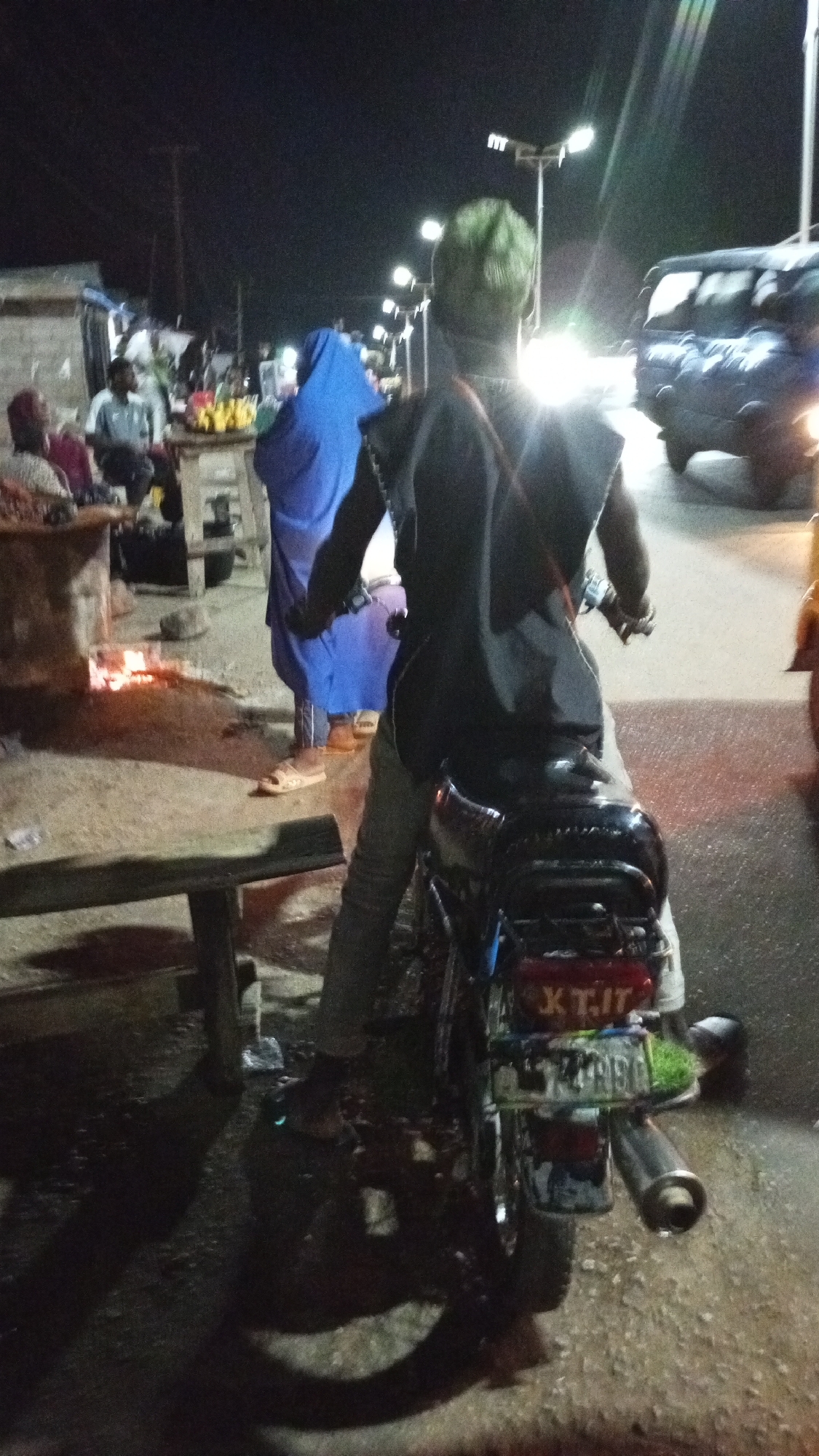 A nighttime scene in Zango-ndaji, Nigeria shows a man on a motorcycle facing away from the viewer. The motorcycle has a license plate with "X T.I.T." visible on the top line and a second, partially obscured line. The man wears a green cap, a dark-colored top, and light-colored pants. In the background, there's a blue-clad figure, a person sitting at a table, and a fire burning on the ground beside a wooden bench. A vehicle's headlights shine brightly in the distance. Streetlights illuminate the scene. A table is visible with bananas on it.