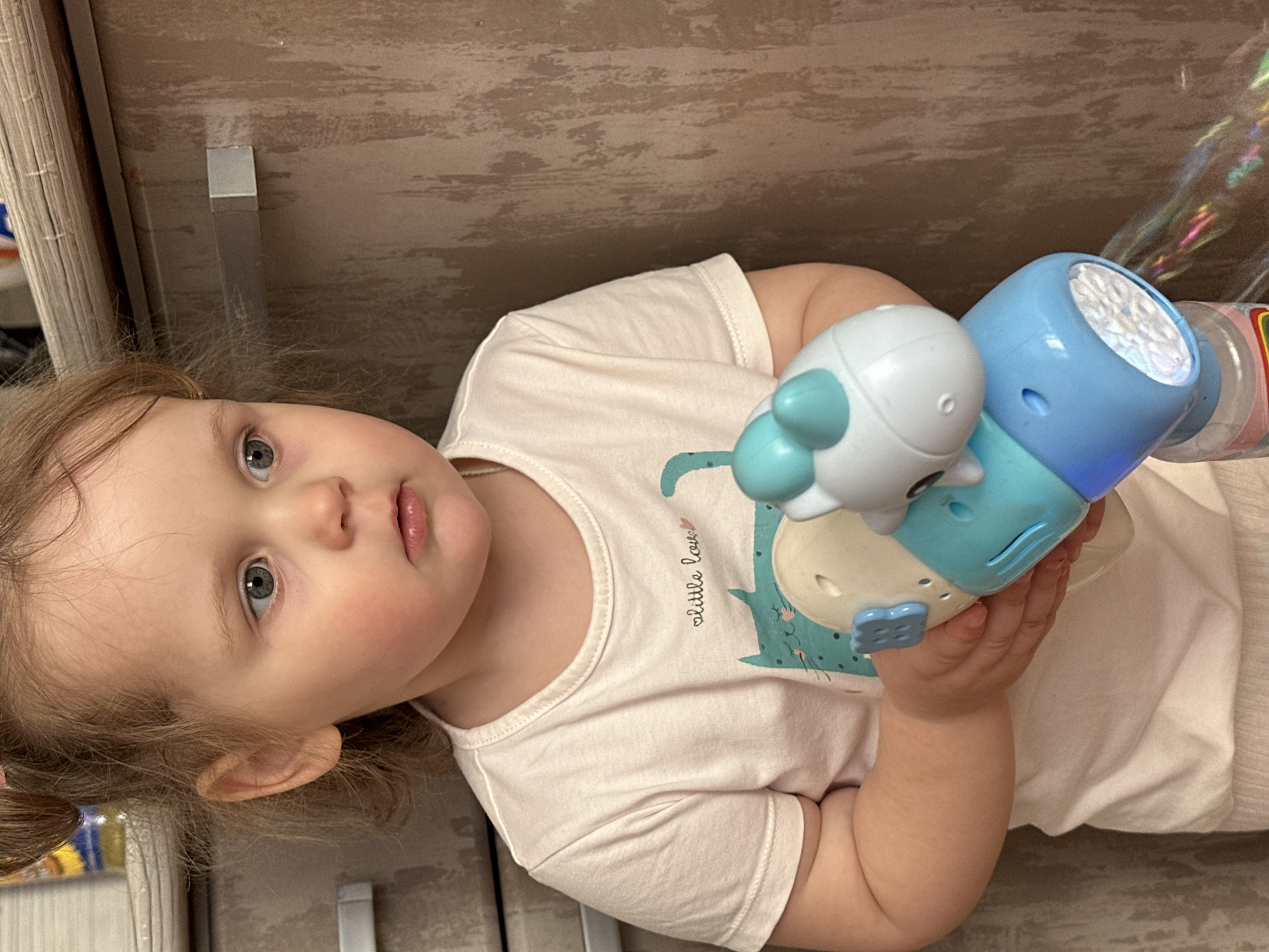 A toddler with fair skin and light hair pulled back into a small ponytail is lying on their side, holding a toy with white, light blue, and blue components. The toy appears to have a cylindrical part with a textured surface. The child is wearing a light-colored t-shirt with a small, green design. The background consists of what seems to be an interior setting with a door or cabinet on the left and a patterned surface behind the child. The image was taken indoors, potentially in a domestic environment, although no clear signs indicate the specific location in Chaikovsky, Russia.