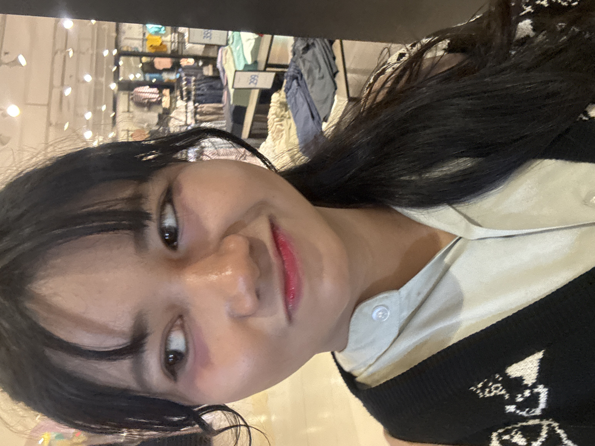 A young woman is taking a selfie in what appears to be a clothing store. She has dark hair, bangs, and is looking to her left. She is wearing a light-colored collared shirt with a dark knitted vest over it. The vest has a white pattern. In the background, there are racks of clothing and store lighting. The store appears to be well-lit with rows of garments displayed on shelves and hanging racks. Prices are visible on some of the displays, suggesting a retail environment.