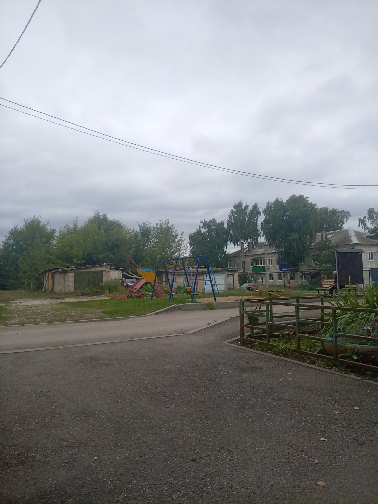The image depicts a residential area in Oktyabrsk, Russia, under an overcast sky. In the foreground, a paved area leads towards a street. To the left, a set of garages constructed from brick and corrugated metal are visible, with a green metal door on one. Adjacent to the garages is a children's playground featuring a blue swing set, a yellow and red slide, and a small red bicycle. Further back, multi-story apartment buildings with light-colored facades and visible windows are situated amongst mature trees. A green metal fence encloses a planted area with vegetation in the lower right portion of the image. The overall scene is one of a modest, somewhat aged urban environment.