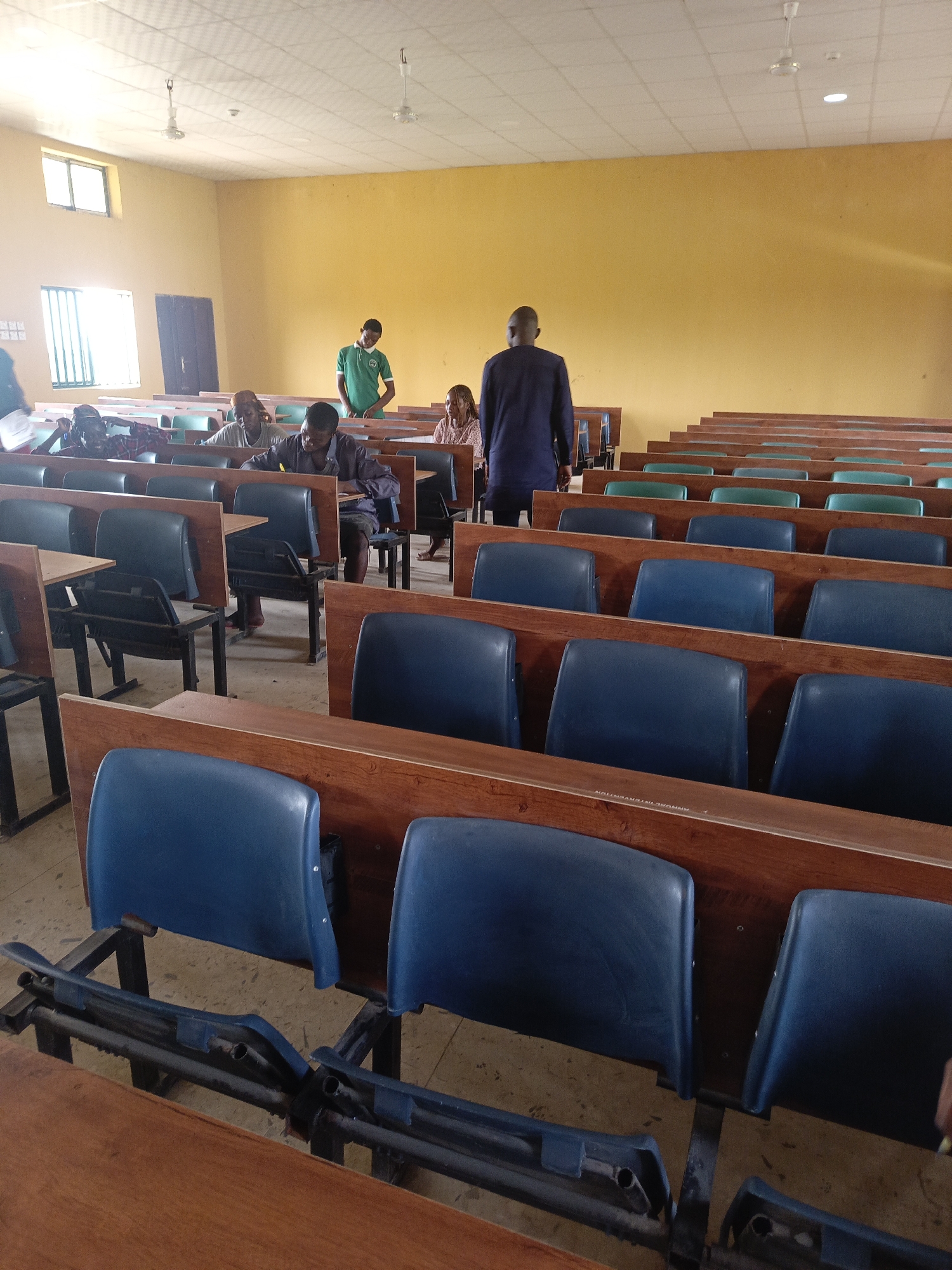 An indoor scene depicts a lecture hall or auditorium, likely at an educational institution in Akwanga, Nigeria. The room is furnished with rows of wooden desks and attached blue and green plastic chairs, many of which are empty. Natural light streams in through two windows on the left wall, complementing the artificial light from ceiling fans and recessed fixtures on the white drop-ceiling. The walls are painted a vibrant yellow.
Several individuals are present. In the background, a man in a dark blue tunic stands with his back to the camera, facing the front of the room. Near the front rows of seated students, a young man in a green polo shirt and a face mask (possibly lowered to his chin) stands, looking towards the students. In the mid-ground, several students are seated at desks. One man in a plaid shirt and another in a dark shirt appear to be writing or looking down at their desks, while a third person in a light-colored shirt is also focused downwards. Further back on the right, a woman with long hair is seated, looking towards the front.
A notable detail in the foreground is a broken chair or footrest, indicating some wear and tear on the furniture. On one of the wooden desks in the middle foreground, faint text "ANNUAL INTERVENTION" is visible. The scene suggests a class, lecture, or possibly an examination in progress or about to commence, with students engaged in their tasks and staff members present.