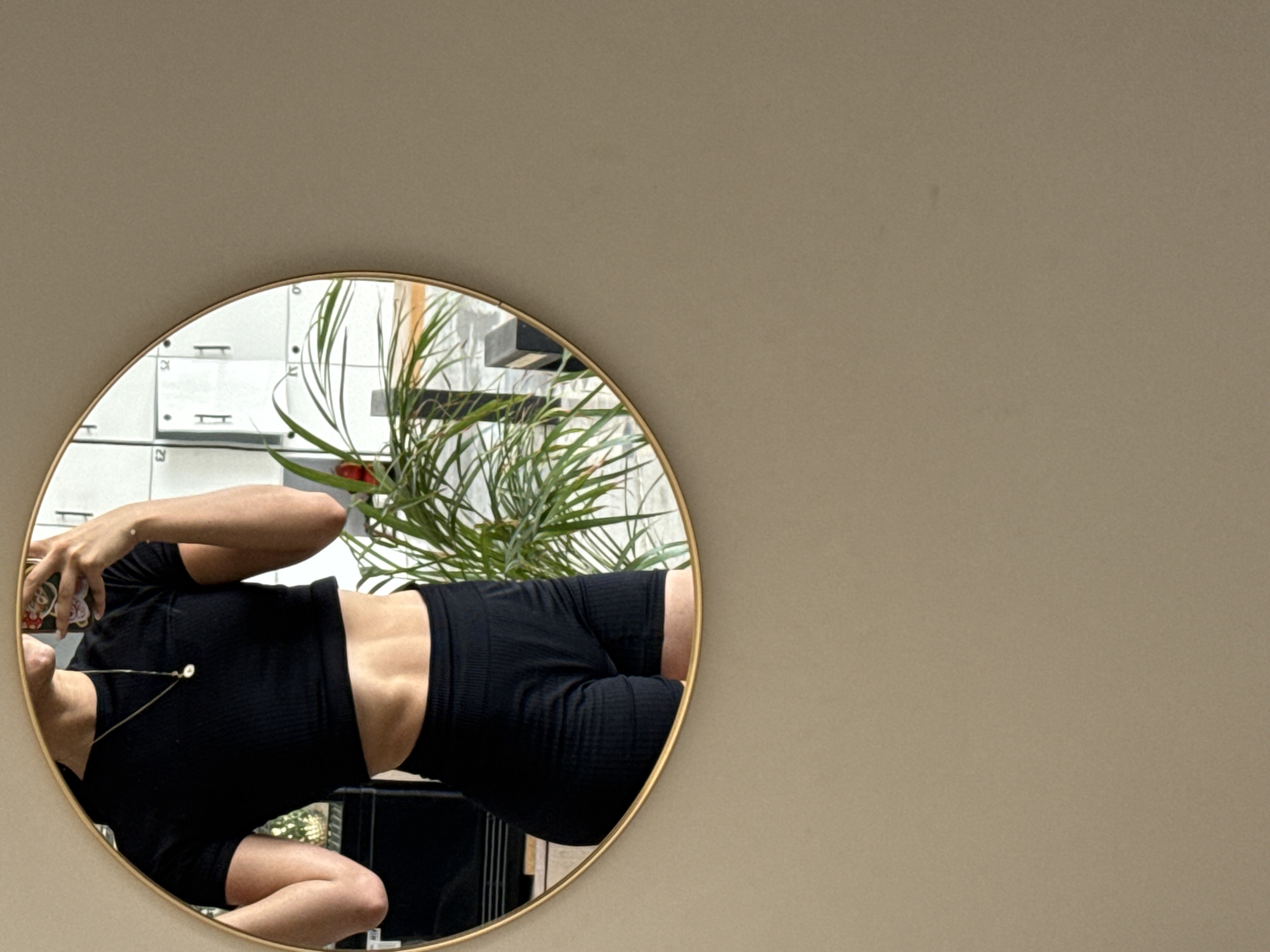 A photograph shows a reflection in a round mirror with a thin gold frame. The mirror is situated against a solid taupe or light brown wall with no visible texture or detail. Within the mirror's reflection, a woman dressed in a black ribbed crop top and matching black ribbed shorts is seen holding a mobile phone in her left hand, presumably taking the picture. Her face is partially obscured by the phone. The top reveals part of her abdomen. Behind her, in the reflection, is a plant with long, thin green leaves and a red flower, with what appears to be a set of locker doors or storage compartments with numerals on them, presumably to facilitate package storage or retrieval. A countertop with some items is also partially visible in the reflection. The location is inferred to be in the city of Puebla, Mexico, based on potential clues and hints.
