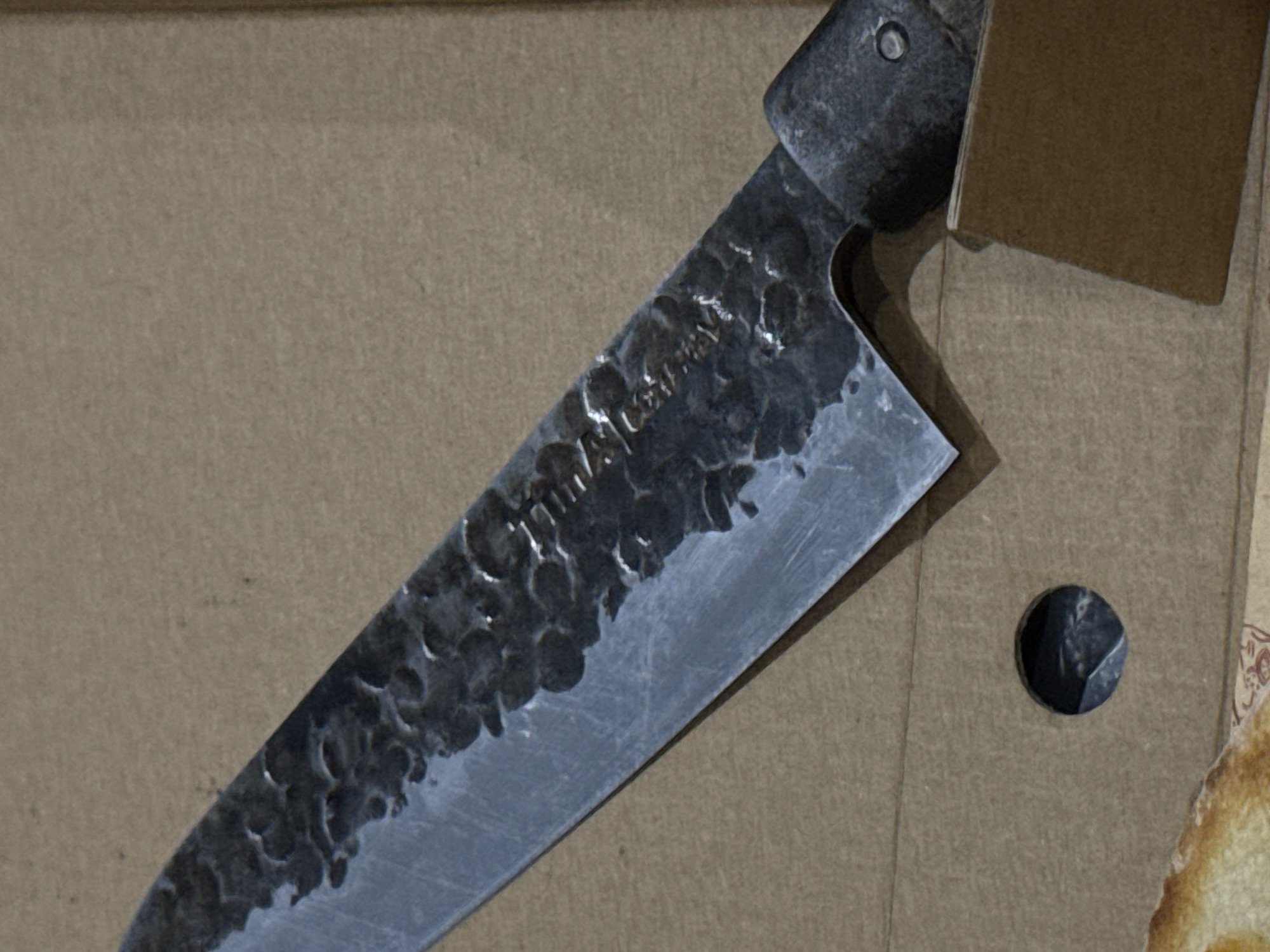 A close-up shot shows a knife with a hammered steel blade resting on a light brown, possibly cardboard, surface. The knife's blade has visible textured indentations. Part of the knife's handle is also visible, along with a portion of a brown box or packaging behind the blade. A circular opening reveals a small, dark-colored object inside. There is no indication of Đà Nẵng, Vietnam in the image.