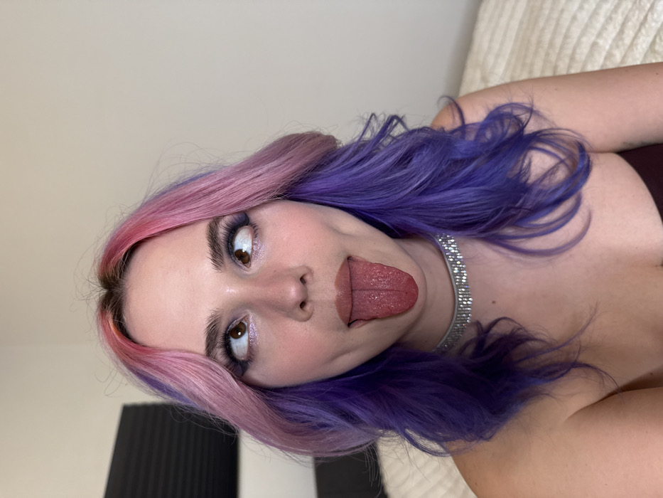 A female subject is depicted in a close-up indoor portrait, visible from the chest up. She has long, wavy hair featuring prominent pink and purple dye, with darker roots visible at the scalp. Her eyes are crossed and directed upwards, while her tongue is extended downwards from her mouth. She wears a multi-row silver-toned rhinestone choker necklace and a dark-colored, possibly strapless, garment. Facial makeup includes dark eyeliner, emphasized eyelashes, and a light lip color. The background consists of a plain, light-colored wall. A dark, pleated object is partially visible on the left side, and a light-colored, textured fabric, consistent with bedding, is present on the right side. No identifying landmarks or specific architectural features are present in the image to determine the precise location within the City of Zagreb, Croatia.