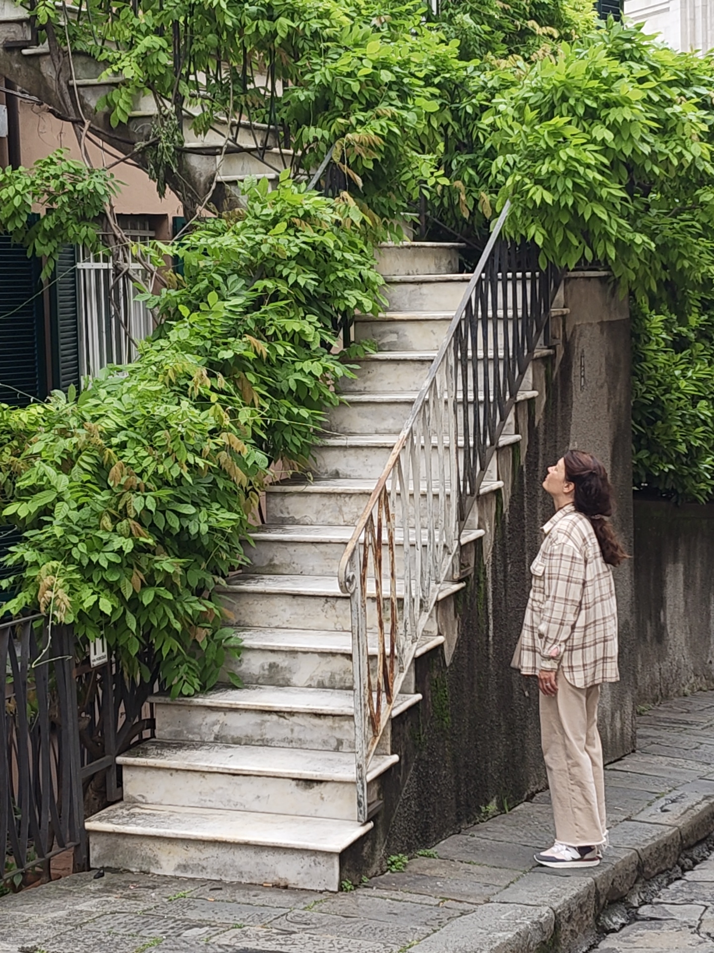 A woman with long brown hair stands on a cobblestone street, looking up at a marble staircase that is overgrown with lush green foliage. The staircase is flanked by a weathered wall on one side and a decorative metal railing on the other. A portion of a building with window shutters is visible behind the greenery to the left of the stairs. The scene is captured in natural light, suggesting an outdoor urban setting. The location is in Genoa, Italy.