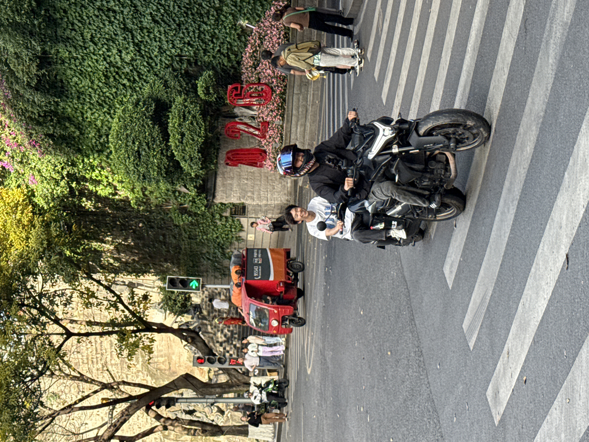 The image depicts a street scene in Yuzhong District, China, featuring a motorcycle with two riders on a crosswalk. The driver is wearing a black jacket and a helmet, while the passenger is wearing a white t-shirt. The motorcycle is black and white. Pedestrians are on the crosswalk near the motorcycle. A red three-wheeled vehicle is also visible. A green traffic light is lit for crossing traffic, and red traffic light is lit for traffic in the foreground. A building with decorative floral arrangements and the numbers "1921" is in the background. The environment includes trees and other vegetation.