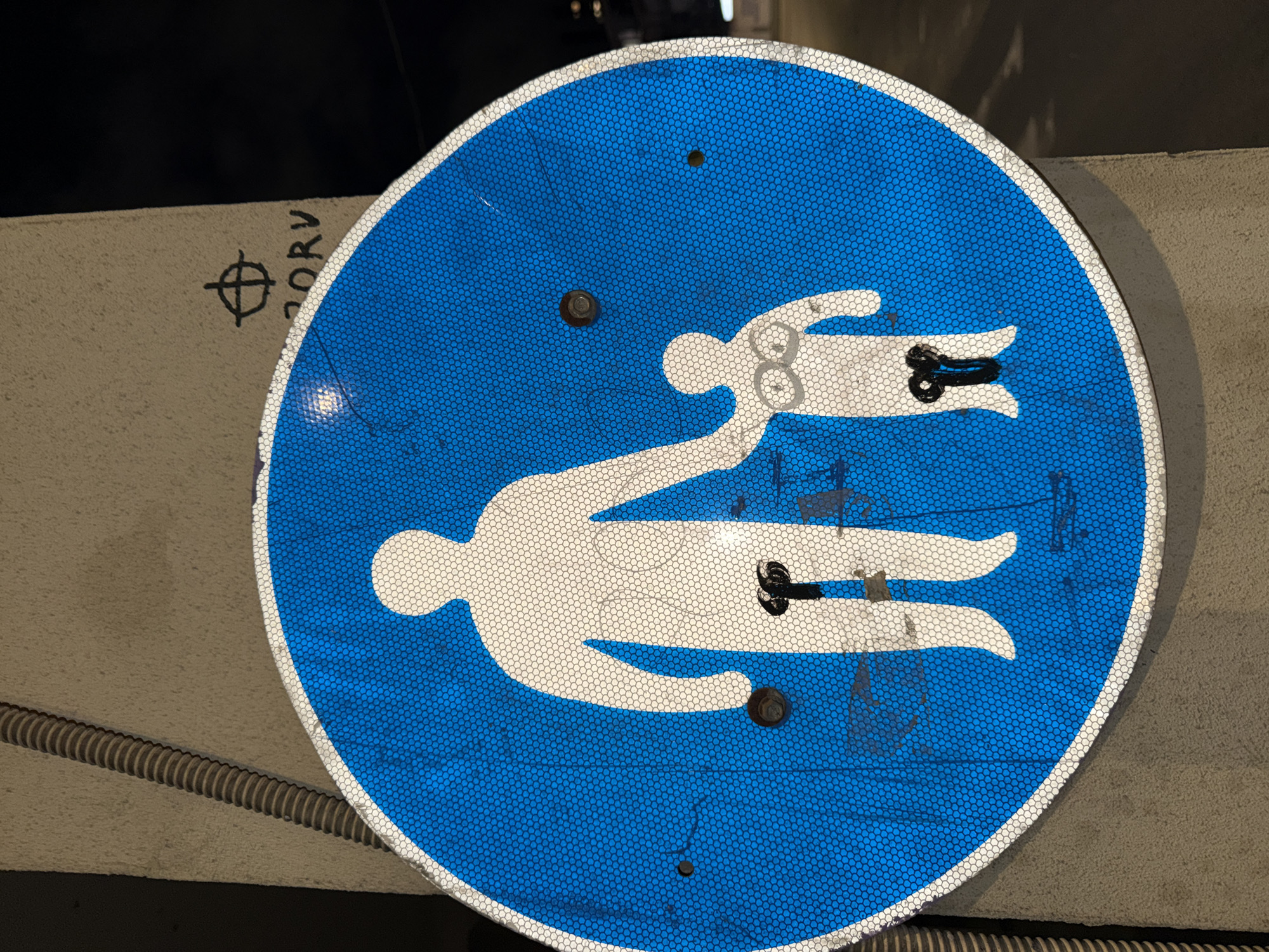 A circular blue pedestrian sign, approximately 60-70 cm in diameter, is affixed to a textured beige concrete pillar in Tel-Aviv, Israel. The sign features white silhouettes of an adult figure holding the hand of a smaller child figure, indicating a pedestrian path. The sign's surface is reflective with a visible hexagonal honeycomb pattern.
The white silhouettes have been modified:
* The adult figure has a black, phallic-shaped object, possibly a plastic or rubber toy or a cut-out, affixed to its groin area, hanging downwards.
* The child figure has two small, white, circular objects, resembling eyes or breasts, drawn or affixed to its chest, and a similar black, phallic-shaped object attached to its groin area, also hanging downwards.
The sign is mounted by at least two visible bolts; one near the adult figure's elbow and another near the child figure's left side. Several other small holes are present on the sign, indicative of prior mountings or damage.
To the upper left of the sign, a vertically oriented corrugated gray conduit is visible, running along the concrete pillar. To the upper right, above the sign, black graffiti is present on the concrete, consisting of a circle with a cross inside (resembling the Zodiac Killer symbol) and the letters "ORV" written below it. The background behind the pillar is dark, suggesting a nighttime setting or a dimly lit area.