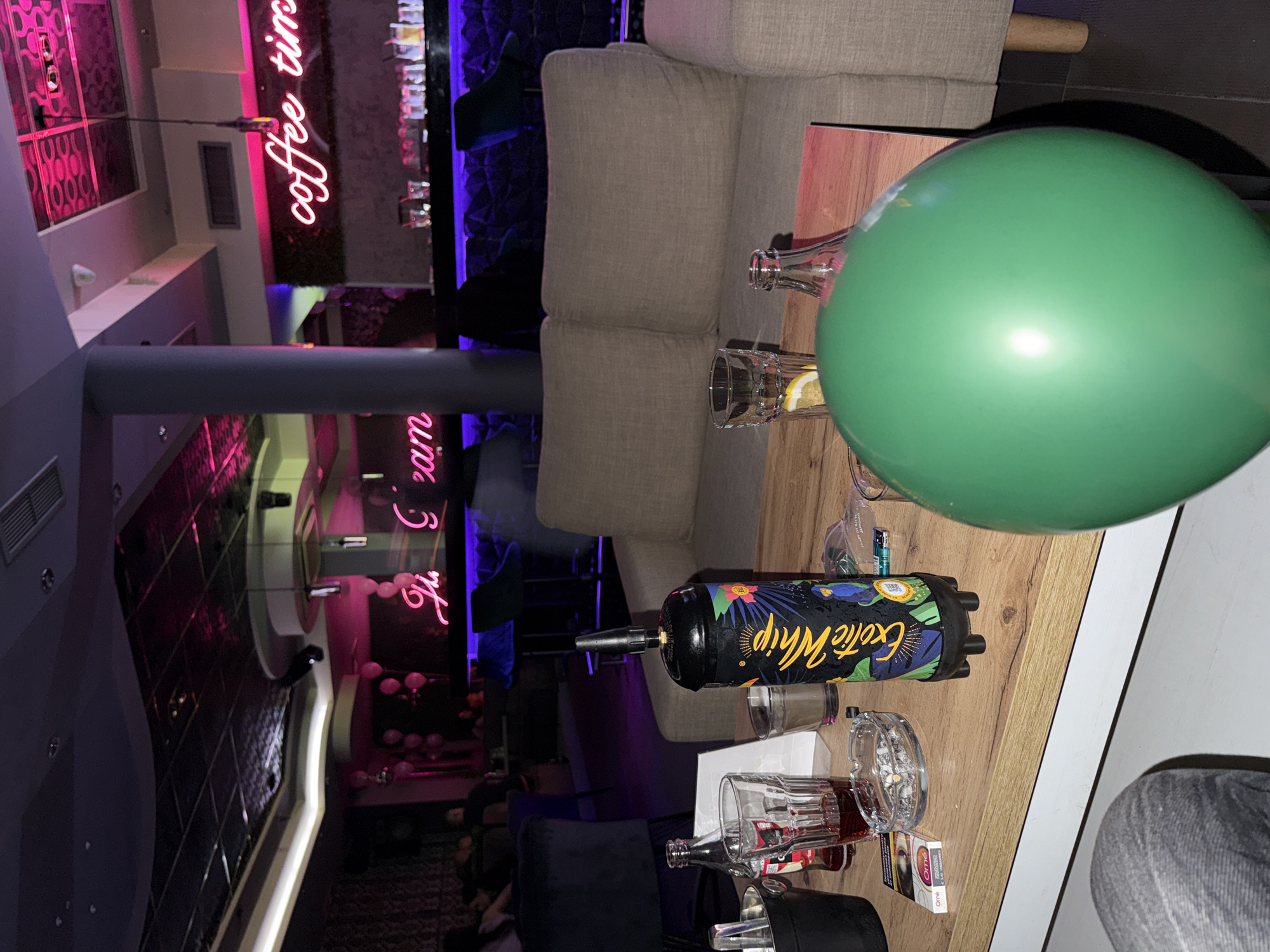 The image shows a lounge or bar setting. There's a seating area with a grey sofa and a wooden table. Various objects are on the table, including glassware (glasses and a bottle), a can of "Exotic Whip" cream charger, an ashtray, and a pack of cigarettes. A green balloon is prominently placed on the table. The background reveals elements of the establishment's decor, including neon signs ("coffee time" and "Ice Cream") and mirrored or reflective surfaces. The setting suggests a modern, possibly upscale, bar or lounge. While I can describe the image in detail, I can not identify the specific location in the city of Karlovo, Bulgaria, without additional information or context.