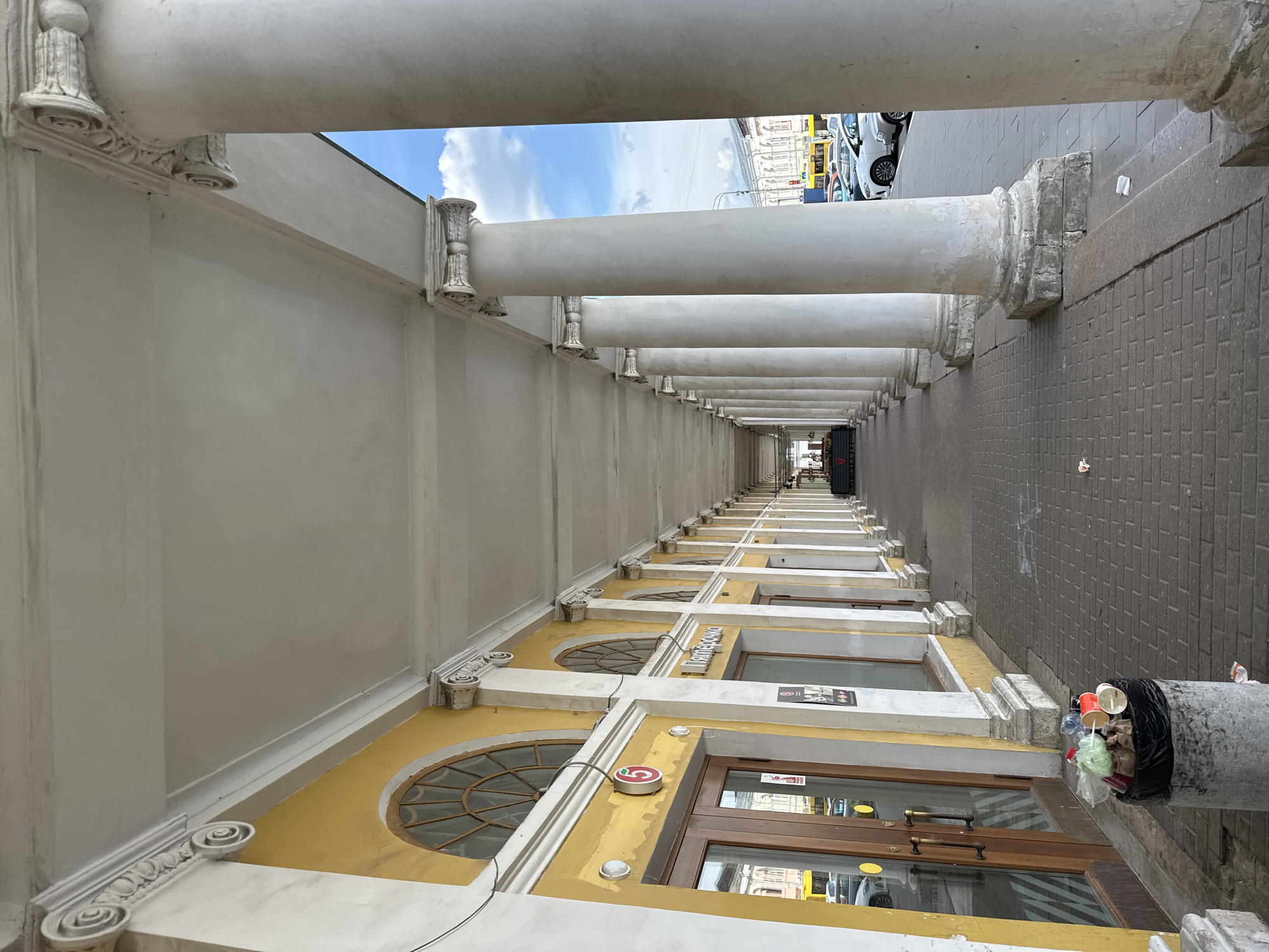 The image shows a perspective view of a covered walkway supported by a colonnade of classical columns. The walkway has a yellow facade on the left, with arched windows and doors. A trash can filled with refuse is visible near the foreground on the left. The ground is paved with interlocking bricks. In the distance, cars and buildings are visible. The sky above is blue with scattered clouds. The scene appears to be a commercial or public space in a city. The sign above one of the doors reads "Пятерочка," a Russian supermarket chain, suggesting the location is likely in Russia. Based on the style of architecture and the presence of the "Пятерочка" store, this scene is likely located in Arkhangelsk, Russia, specifically a shopping arcade or market area. There are no people clearly visible in the image, and the focus is on the architectural elements and the environment.