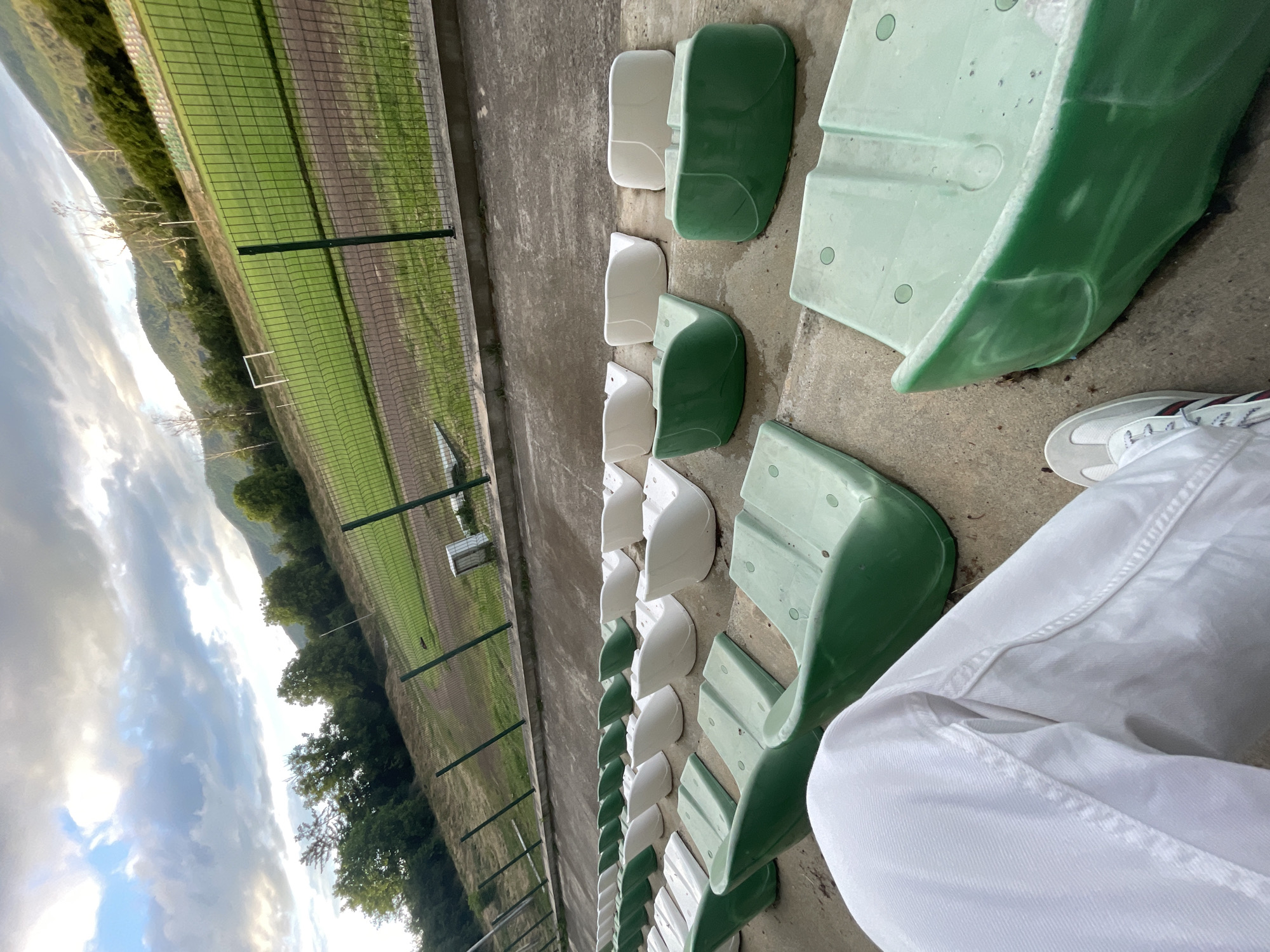 The image displays a quiet outdoor sports facility, likely a small stadium or local sports ground, situated in Ihtiman, Bulgaria. The perspective is from the spectator stands, looking out towards the field.
In the immediate foreground, the lower body of a person is visible, wearing white trousers and white sneakers, indicating they are seated. Ahead of the person, rows of empty stadium seats are arranged on concrete steps. These seats alternate in color, predominantly green and white, with some larger, more robust-looking green seats interspersed with smaller white ones.
Beyond the seating area, a tall green chain-link fence separates the stands from the sports arena. A dirt running track encircles a verdant green grass field, where a small white goalpost can be seen. A line of trees borders the far side of the field. In the distance, undulating hills or mountains rise under a wide, partly cloudy sky, with patches of bright light suggesting diffused sunlight. The lighting indicates it is likely late afternoon or early evening.
No activity or event is taking place on the field, lending a calm and empty atmosphere to the scene. There is no visible text in the image.