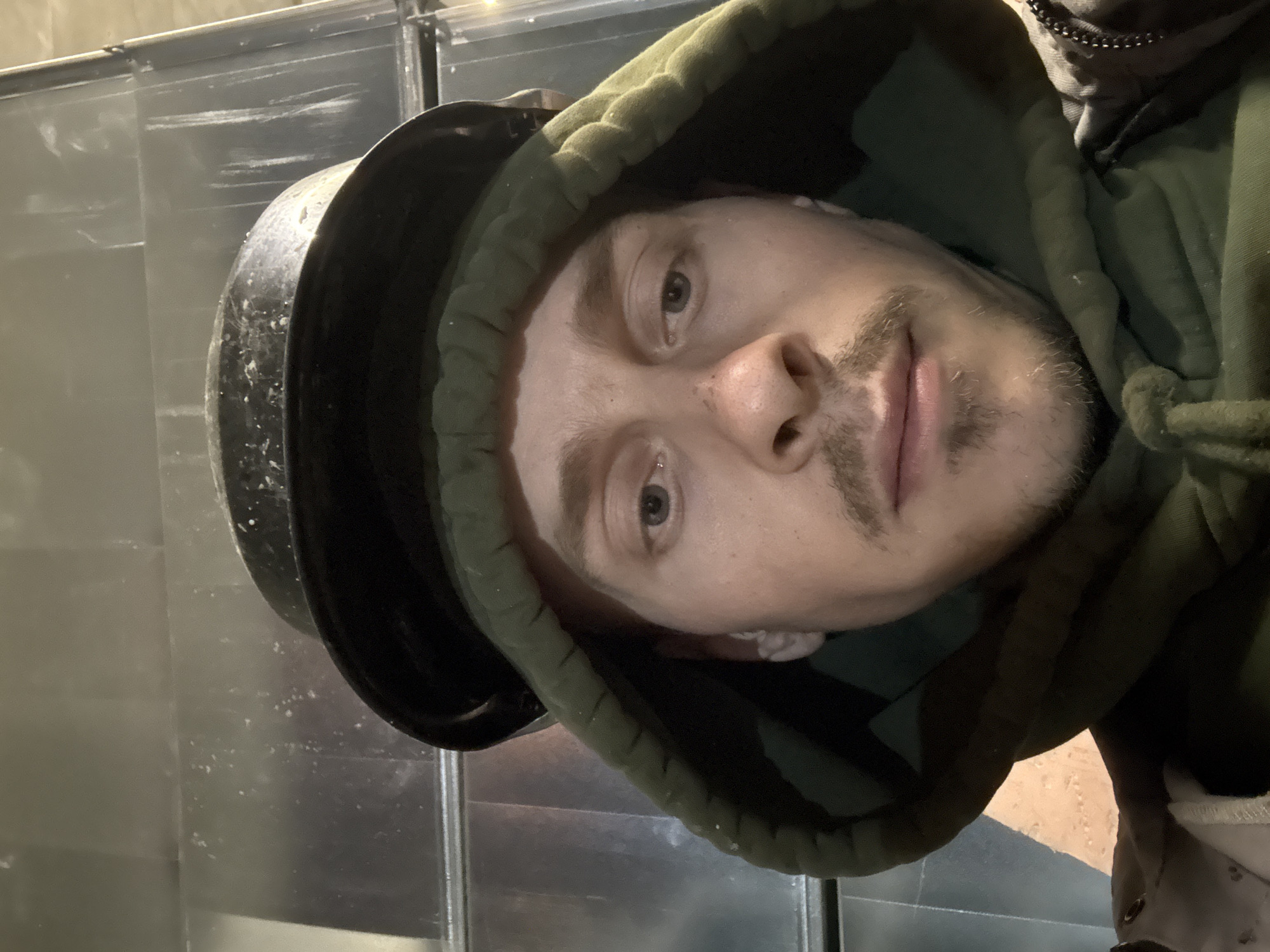 The image is a selfie taken indoors. A young Caucasian male with light skin, blue eyes, and a mustache is wearing a dark pot on his head and a green hooded sweatshirt. The background appears to be a metallic, corrugated wall. The lighting is dim. There is no indication that this image was taken in Moscow, Russia.