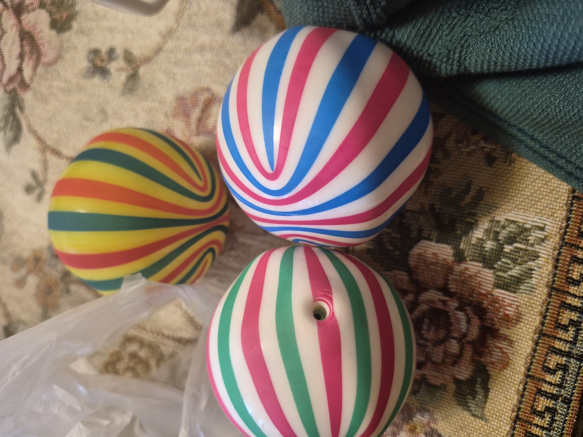The image shows three striped, spherical objects, likely toys or balls, arranged on a patterned fabric surface. One ball, in the foreground to the left, has alternating stripes of white, pink, and green, and features a small circular hole. Another ball, to the right, has alternating white, pink, and blue stripes. A third ball, positioned behind and between the other two, displays stripes of yellow, green, red, and pink. The fabric beneath appears to be a tablecloth or similar textile with a floral and geometric pattern. A portion of a white plastic bag is visible in the upper left of the frame, and a dark green textured fabric is present on the right side. The lighting suggests an indoor setting. The image was captured in Yerevan, Armenia.