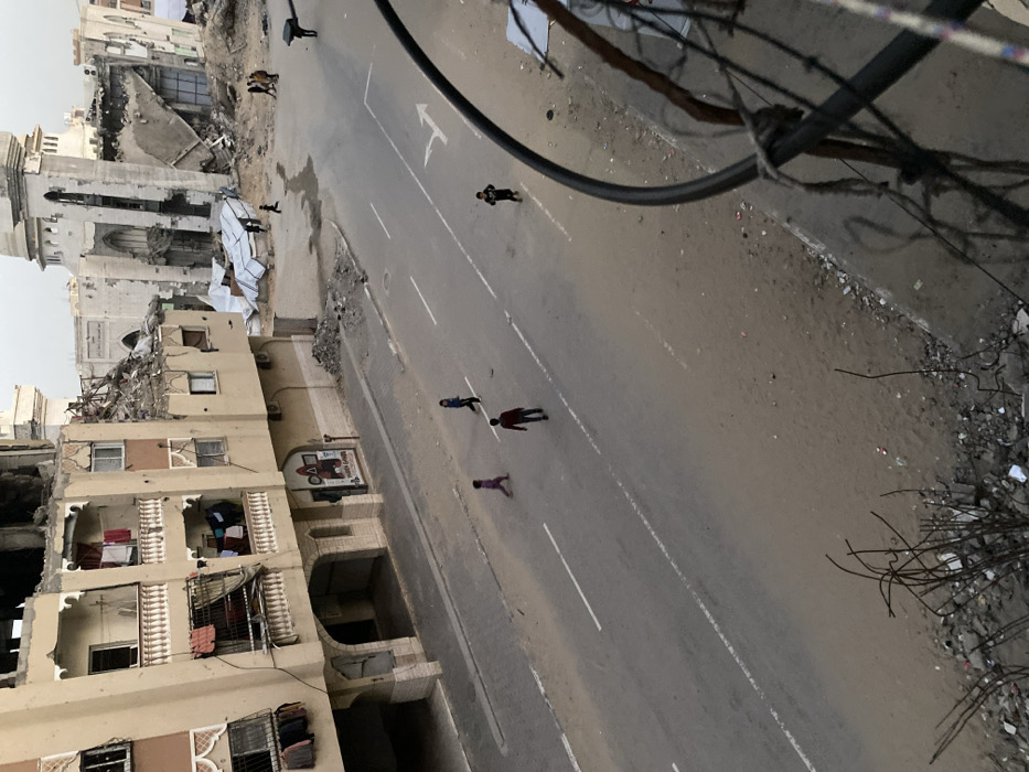 The image captures a street scene in Khan Yunis, Palestinian Territory, characterized by significant structural damage. A wide asphalt road, largely covered in sand and light dirt, runs through the center of the frame, displaying faded white dashed lane markings and a single white right-turn arrow.
On the left side of the street, a multi-story building, light tan in color, exhibits severe damage to its upper sections, with a significant portion of the top floor visibly collapsed and exposing internal structure. Lower floors feature balconies with white balustrades, from which clothes are hanging on lines. A ground-floor shopfront with a distinct red logo is visible.
To the right and further down the street, a large, heavily damaged building, identifiable as a mosque by its architectural style and two minarets, stands partially ruined. Much of its main structure has collapsed into a large pile of rubble, with one minaret still partially standing. White sheeting or tarpaulins are scattered amidst the debris in this area.
On the street itself, four children are observed: one in pink walking left, one in dark clothing walking right, one in dark clothing walking away from the camera, and one in dark clothing (with a white design) running towards the right. Further down the street, near the damaged mosque, a small group of adults or older children are also walking. An individual in dark clothing stands near the right edge of the road, facing away.
The immediate foreground reveals exposed rebar and concrete rubble, indicating the photographer's elevated position from a damaged structure. A thick dark cable and several wires partially obscure the lower right corner of the image. The sky appears overcast or hazy.